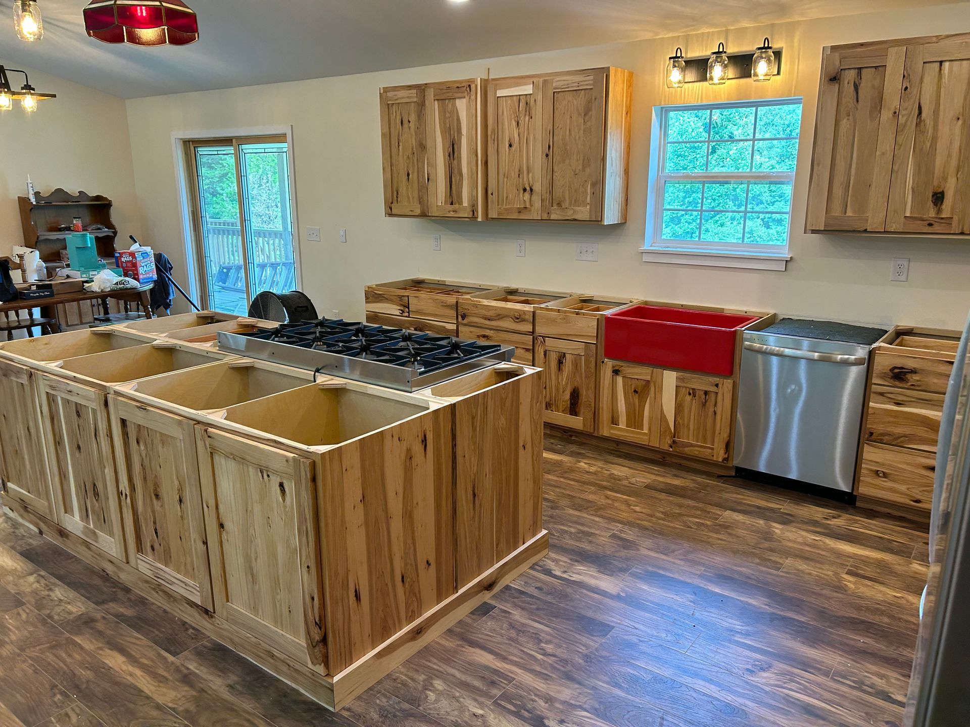 A kitchen undergoing renovation with rustic wooden cabinets, a central island with a cooktop, and a red farmhouse sink.