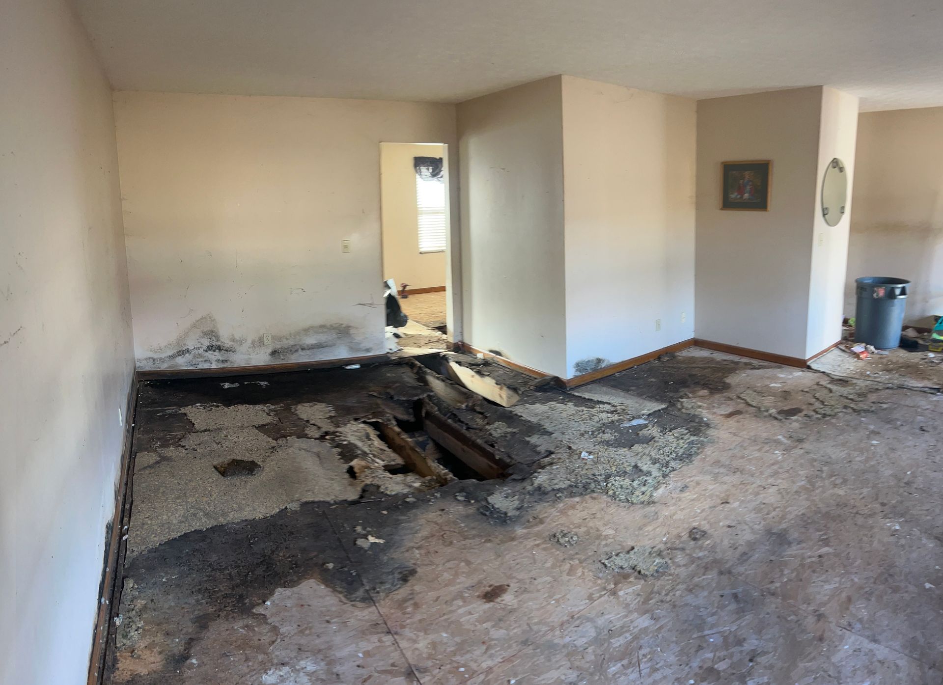 A room with damaged flooring, revealing a dark hole and water-stained walls, suggesting severe interior damage.