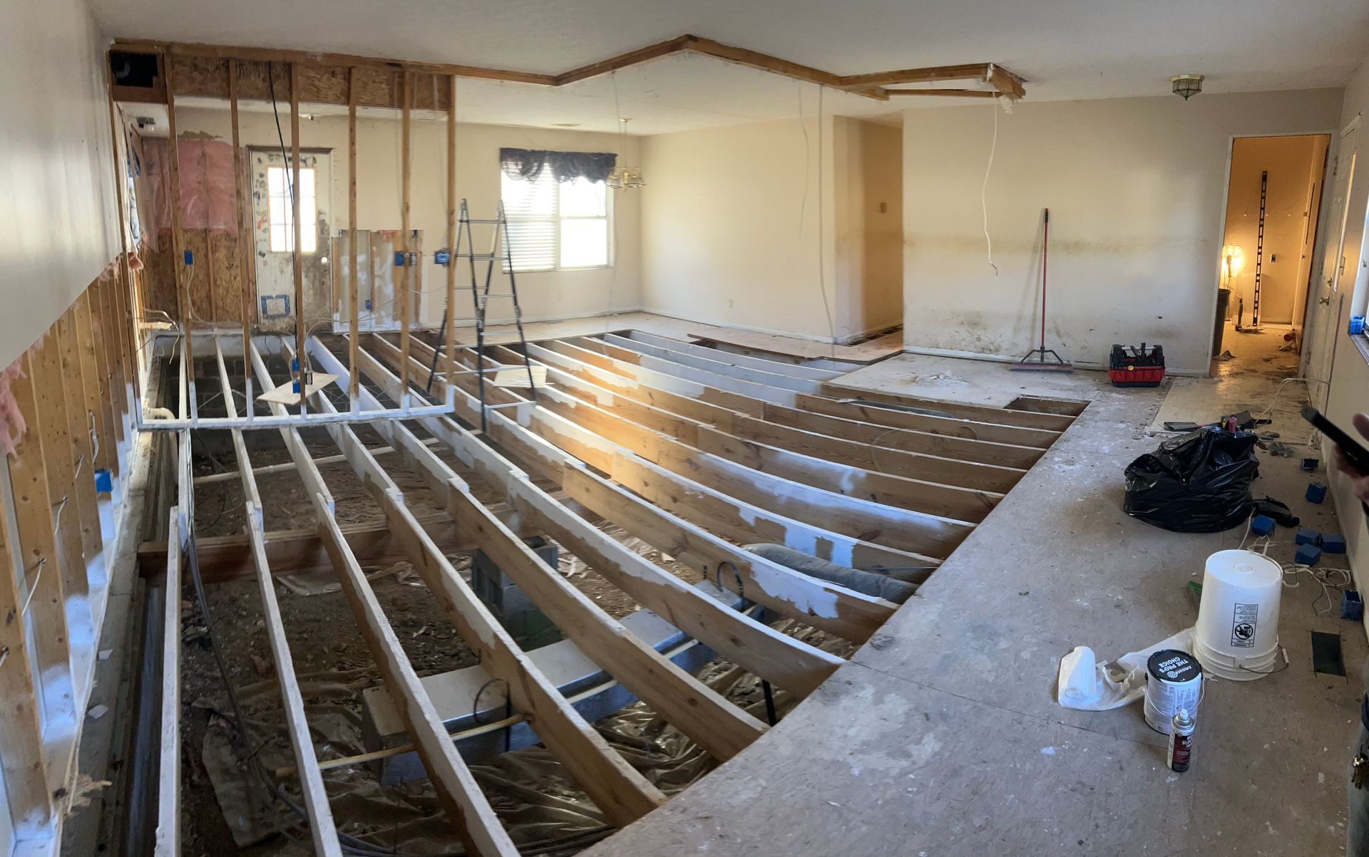 An interior view of a room under renovation, showing exposed floor joists, an open-frame wall, and construction supplies.