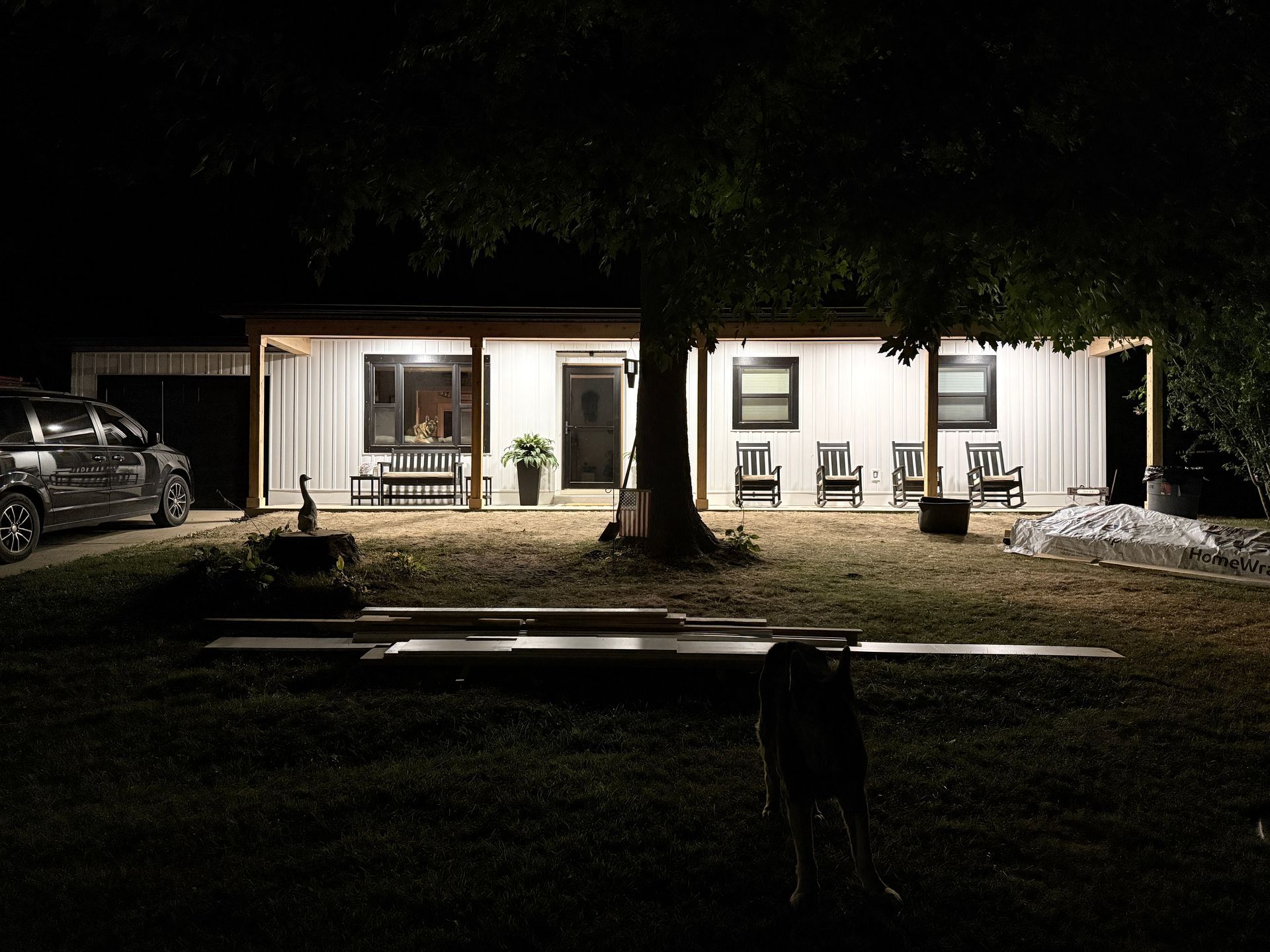 Lit house with porch chairs at night, tree in foreground, car parked on the left, and yard in darkness.