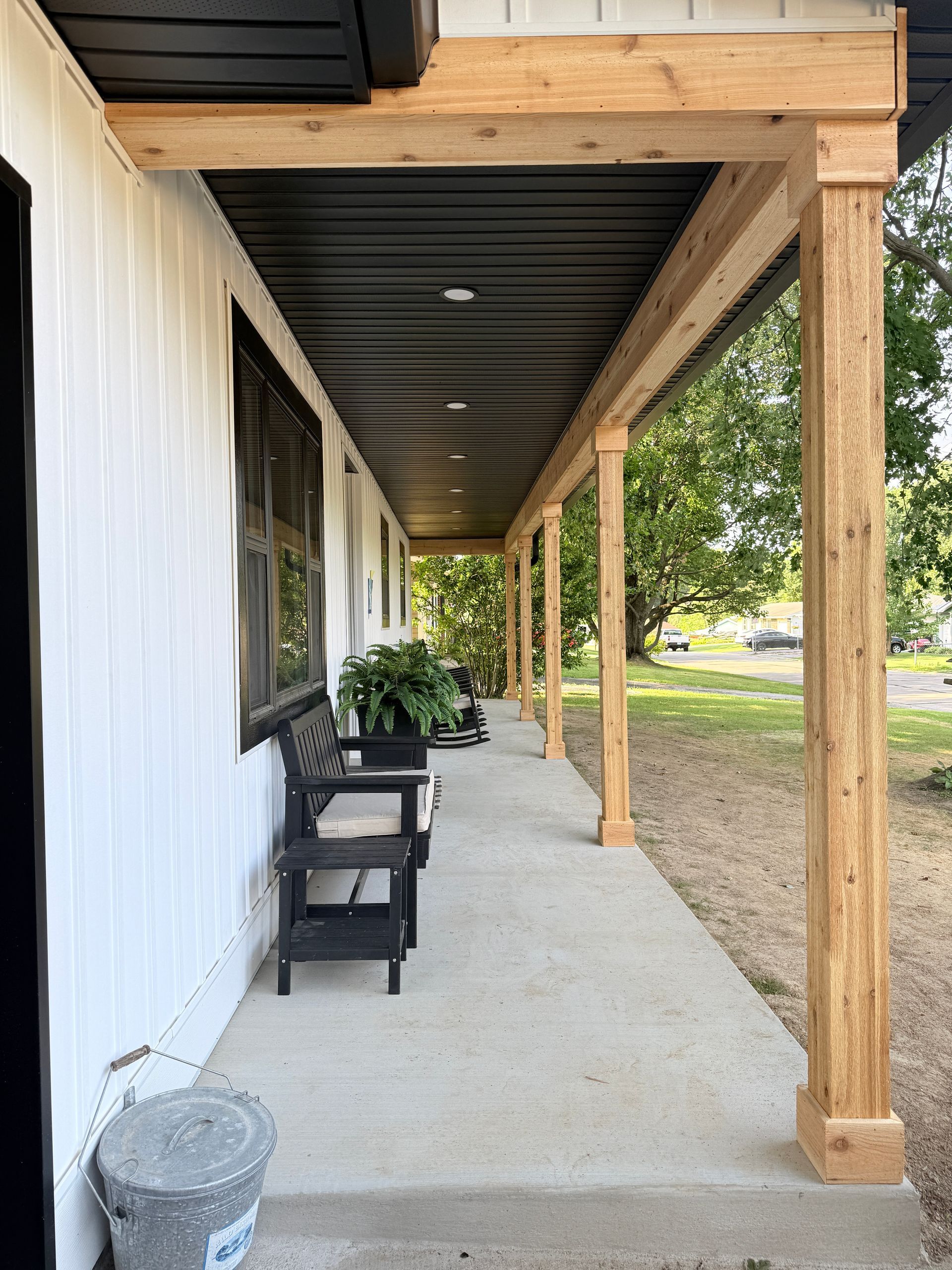 Covered porch with black chairs, a white wall, and wooden posts beside a gravel yard.