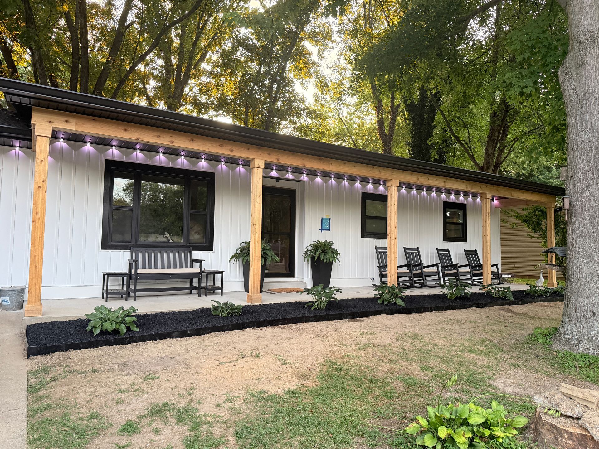 White house with covered porch, black trim, benches, and string lights in a wooded yard