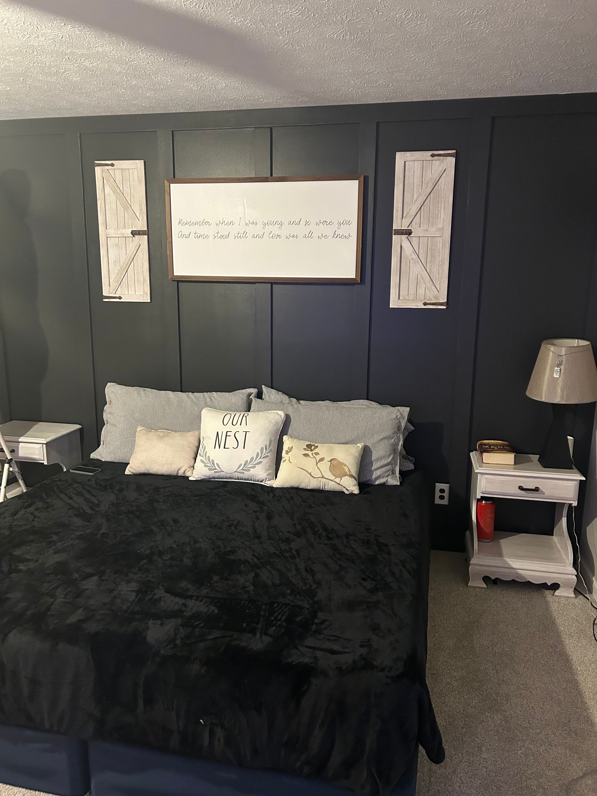 A bedroom with a dark blue board-and-batten wall, a bed with black bedding, white pillows, and a framed sign above it.