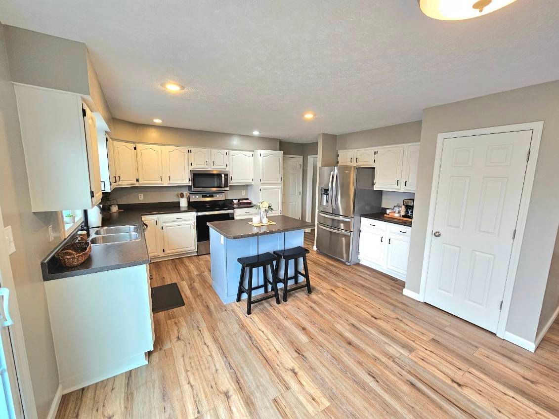 A modern kitchen with white cabinets, a blue island with two black stools, wood-style flooring, and a stainless fridge.