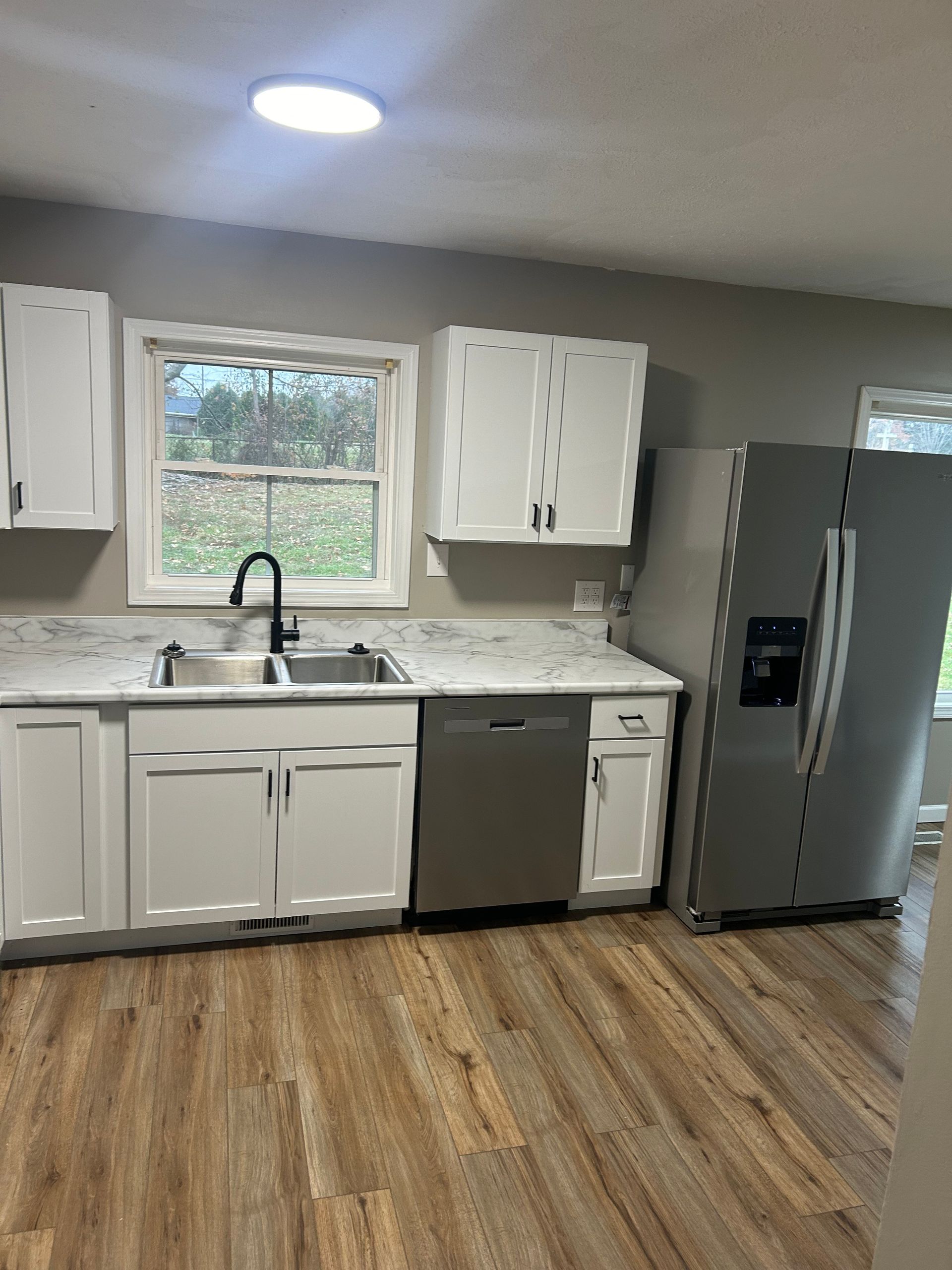 A kitchen with white cabinets, marble countertops, stainless steel appliances, and wood-look flooring under a window.