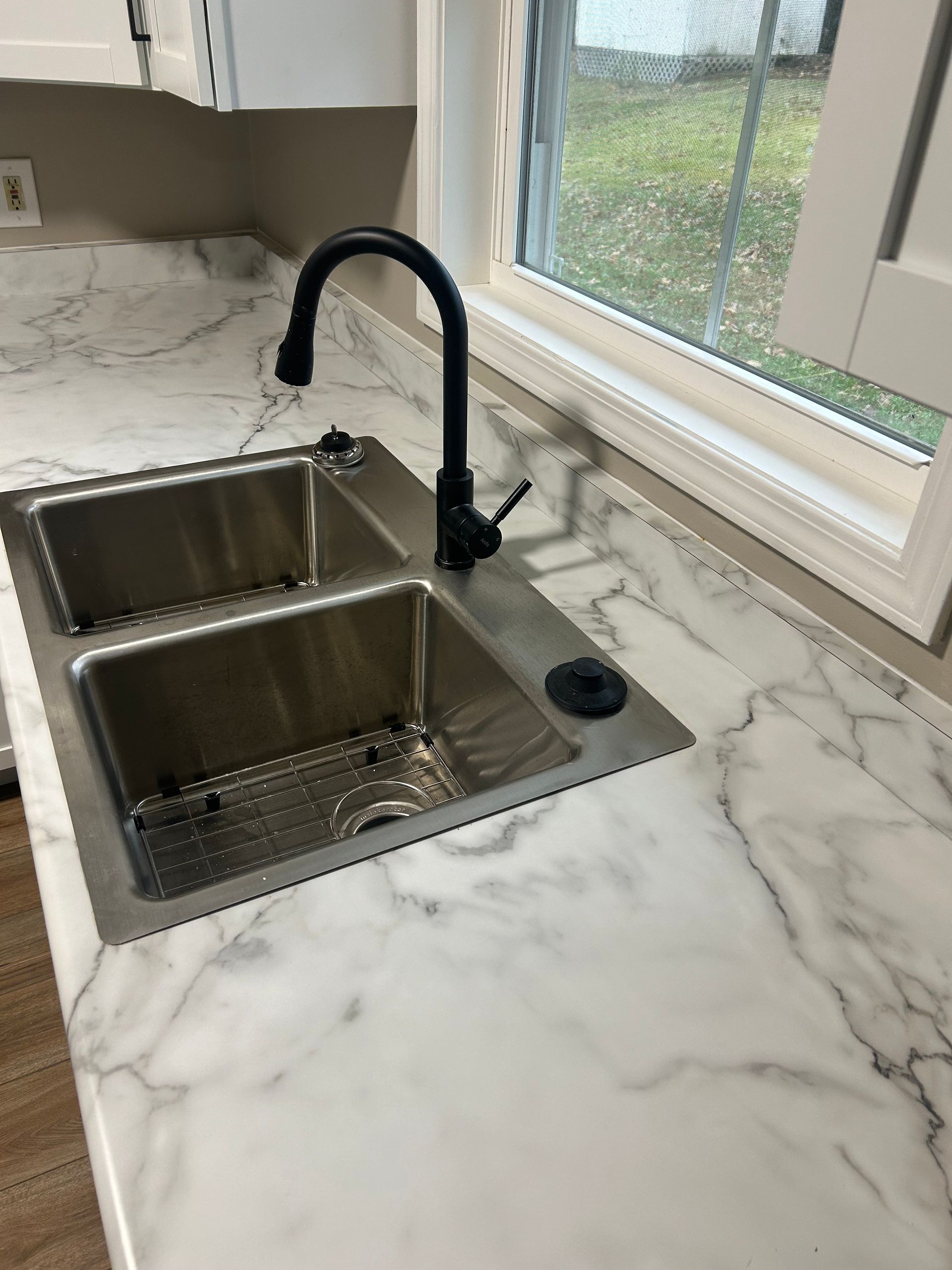 A stainless steel double-basin kitchen sink with a black faucet, set in a white marble-patterned countertop by a window.