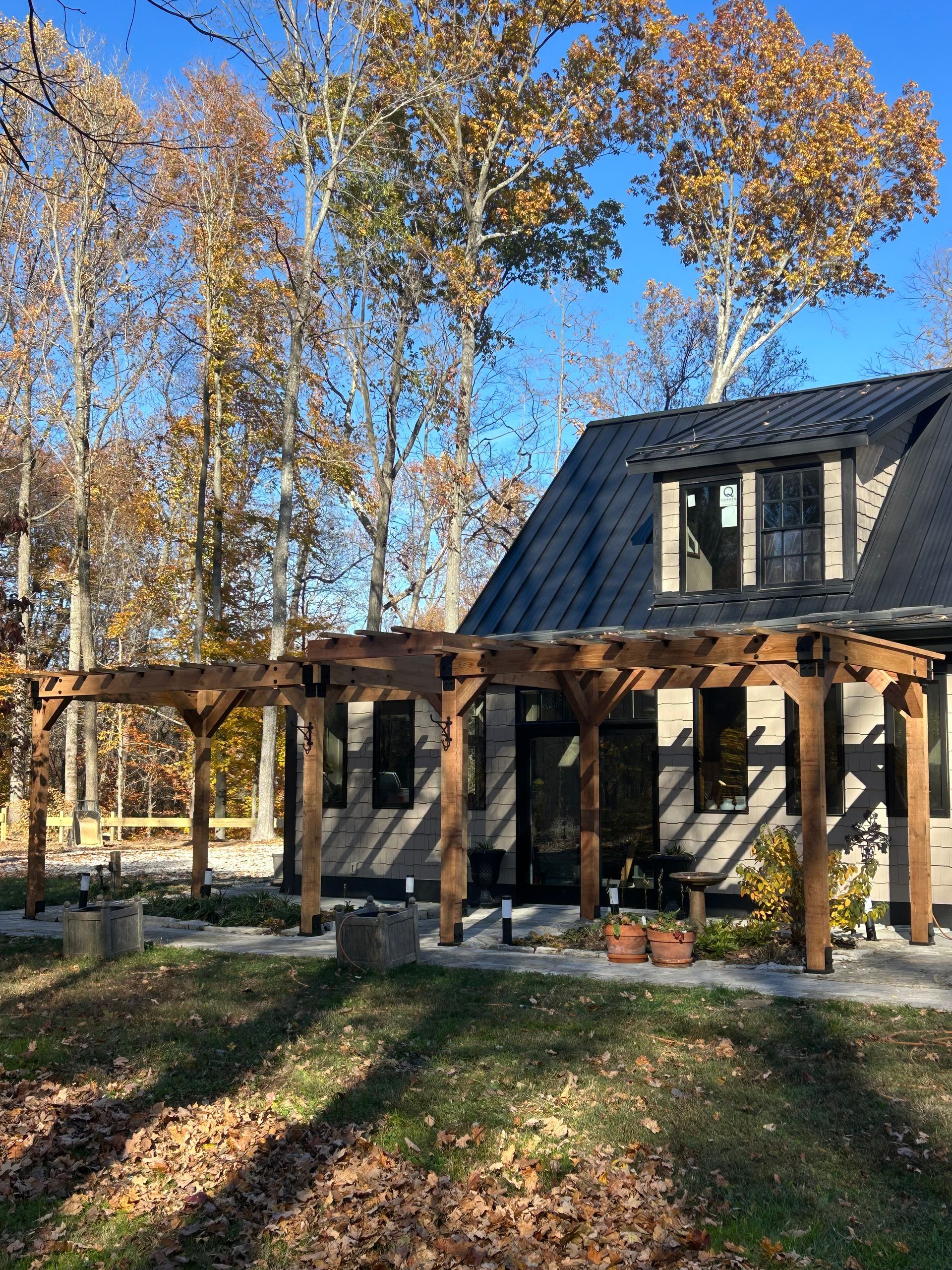 A wooden pergola stands on a stone patio outside a house with a dark metal roof, set against a backdrop of autumn trees.