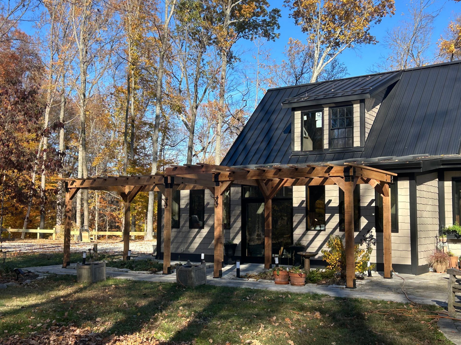 A house with a black metal roof and a wooden pergola over a stone patio, surrounded by trees with autumn foliage.