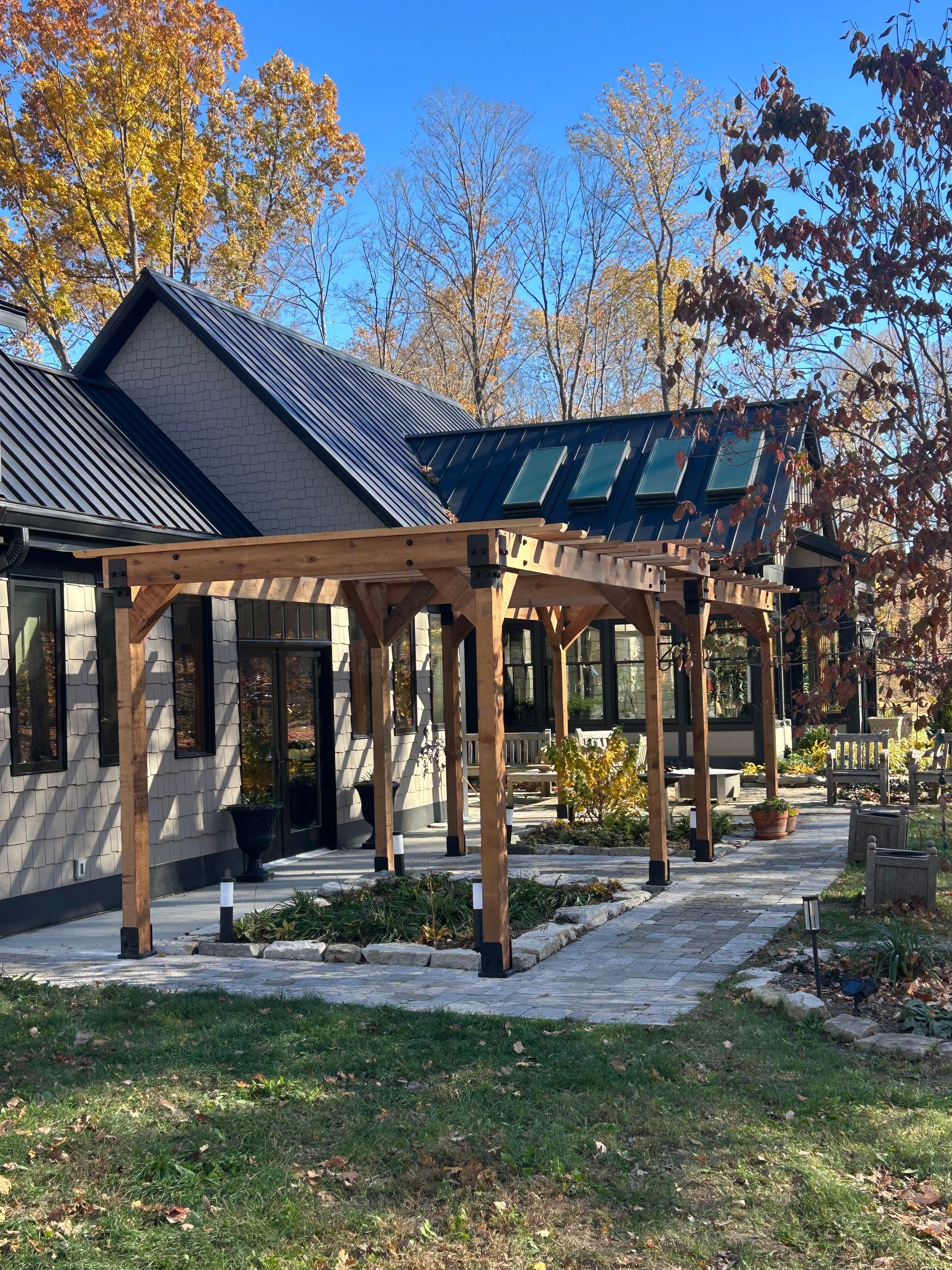 A modern house with a black metal roof and a wooden pergola over a stone patio, surrounded by autumn trees.