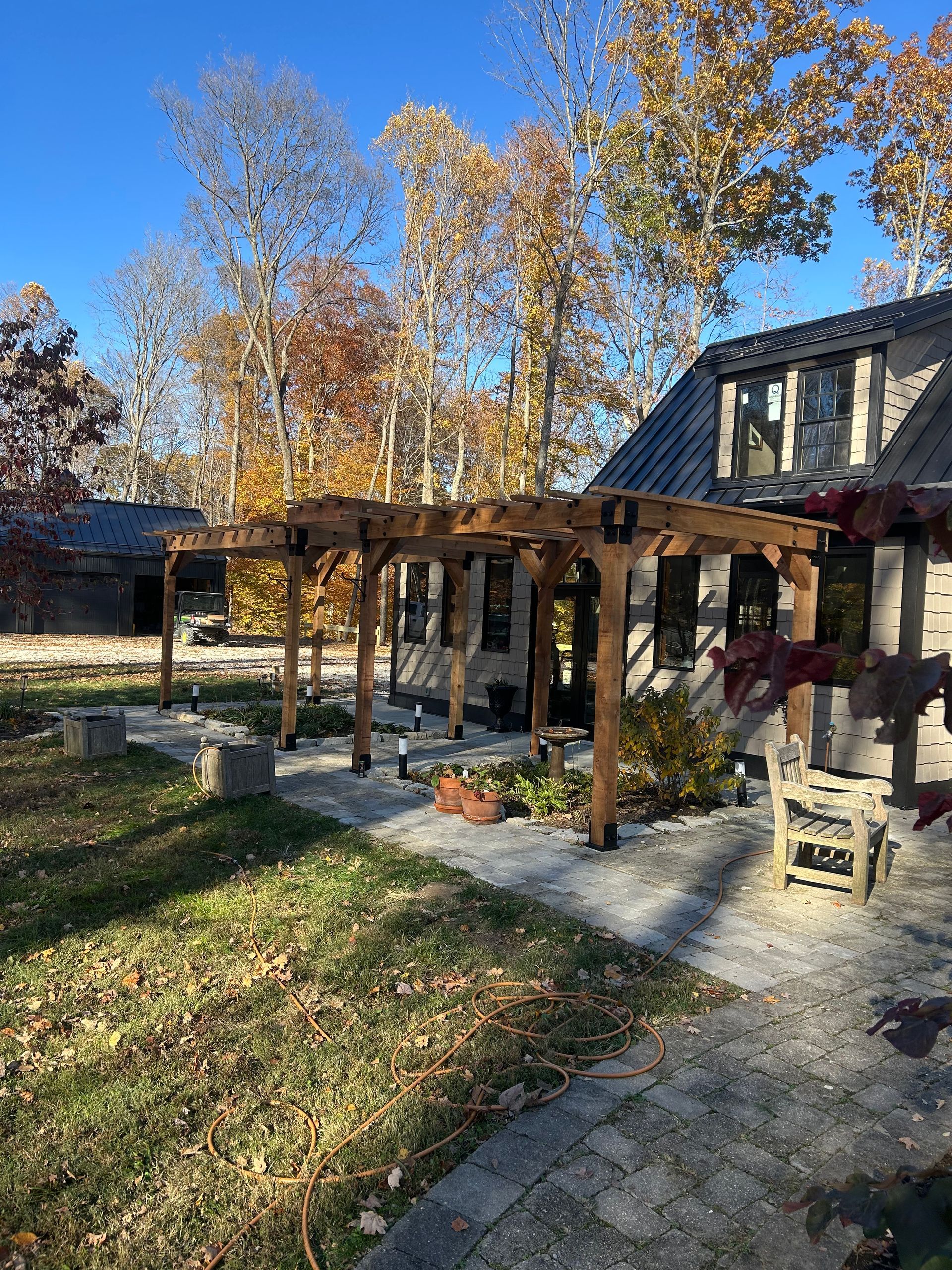 A wooden pergola attached to a stone house on a stone patio, set against a backdrop of trees with autumn foliage.