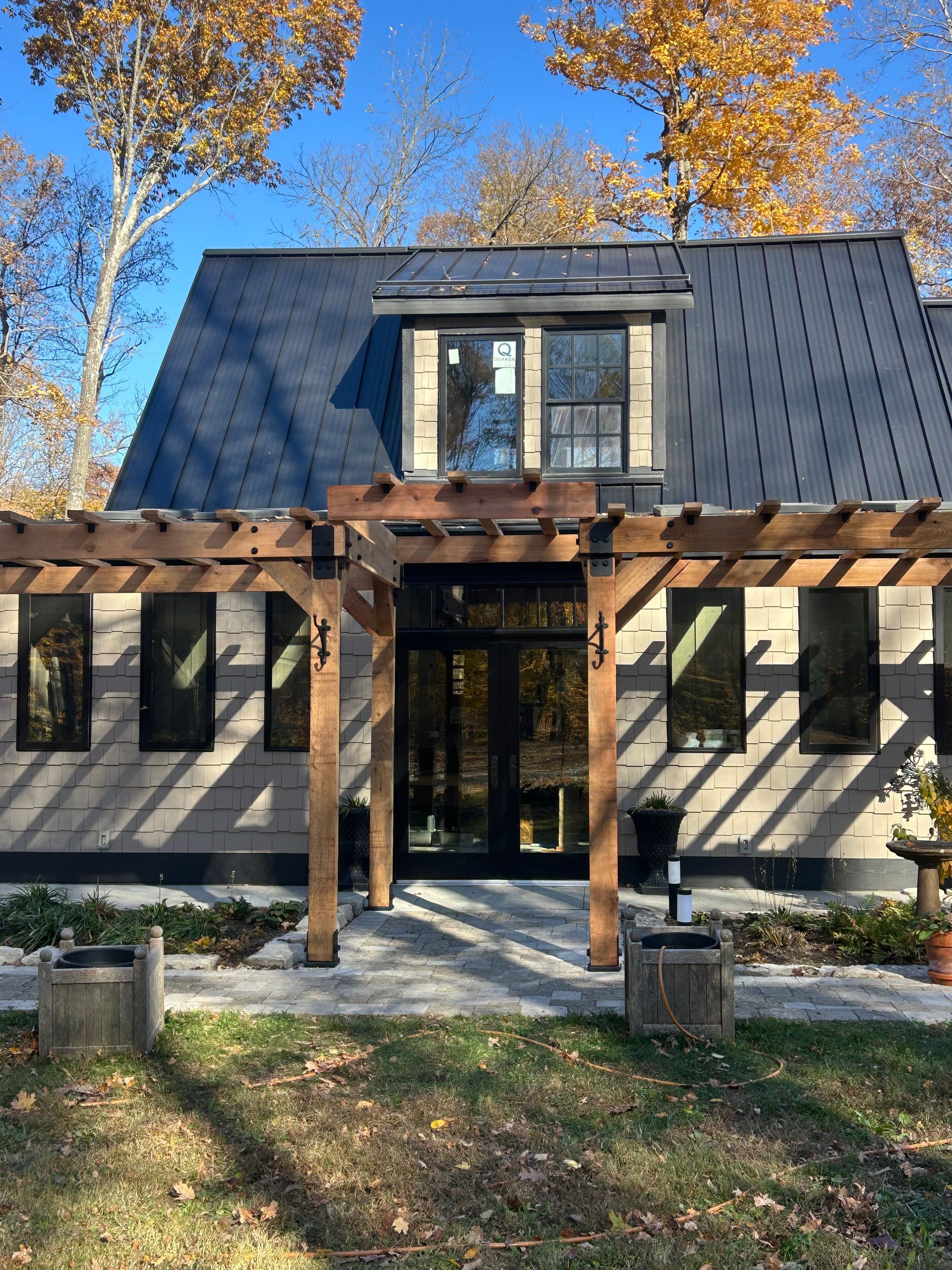 A modern house with a black metal roof, a wooden pergola over the entrance, and trees with autumn leaves in the background.