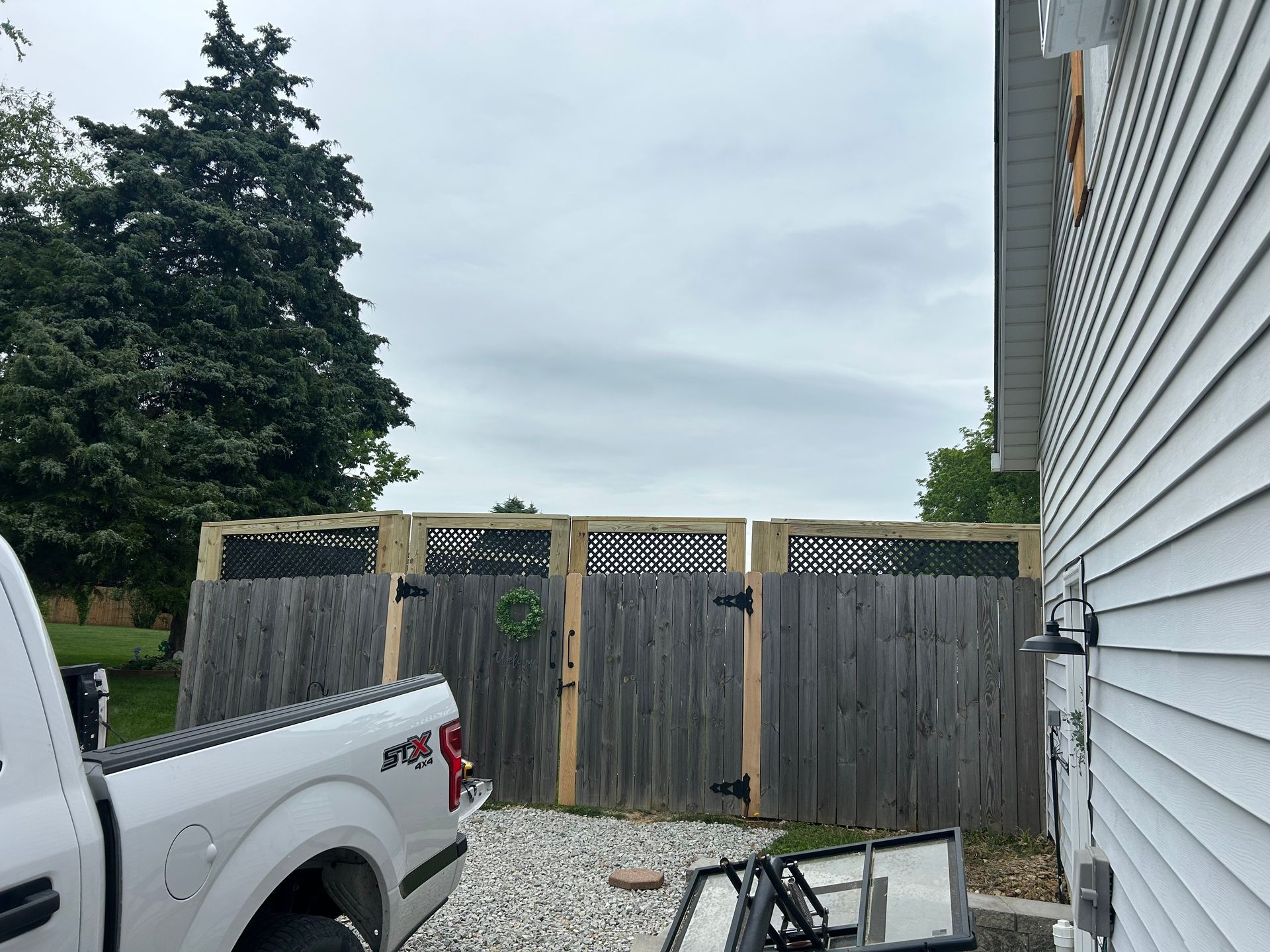 A white pickup truck parked next to a wooden fence with trellis sections on a gravel driveway by a house.