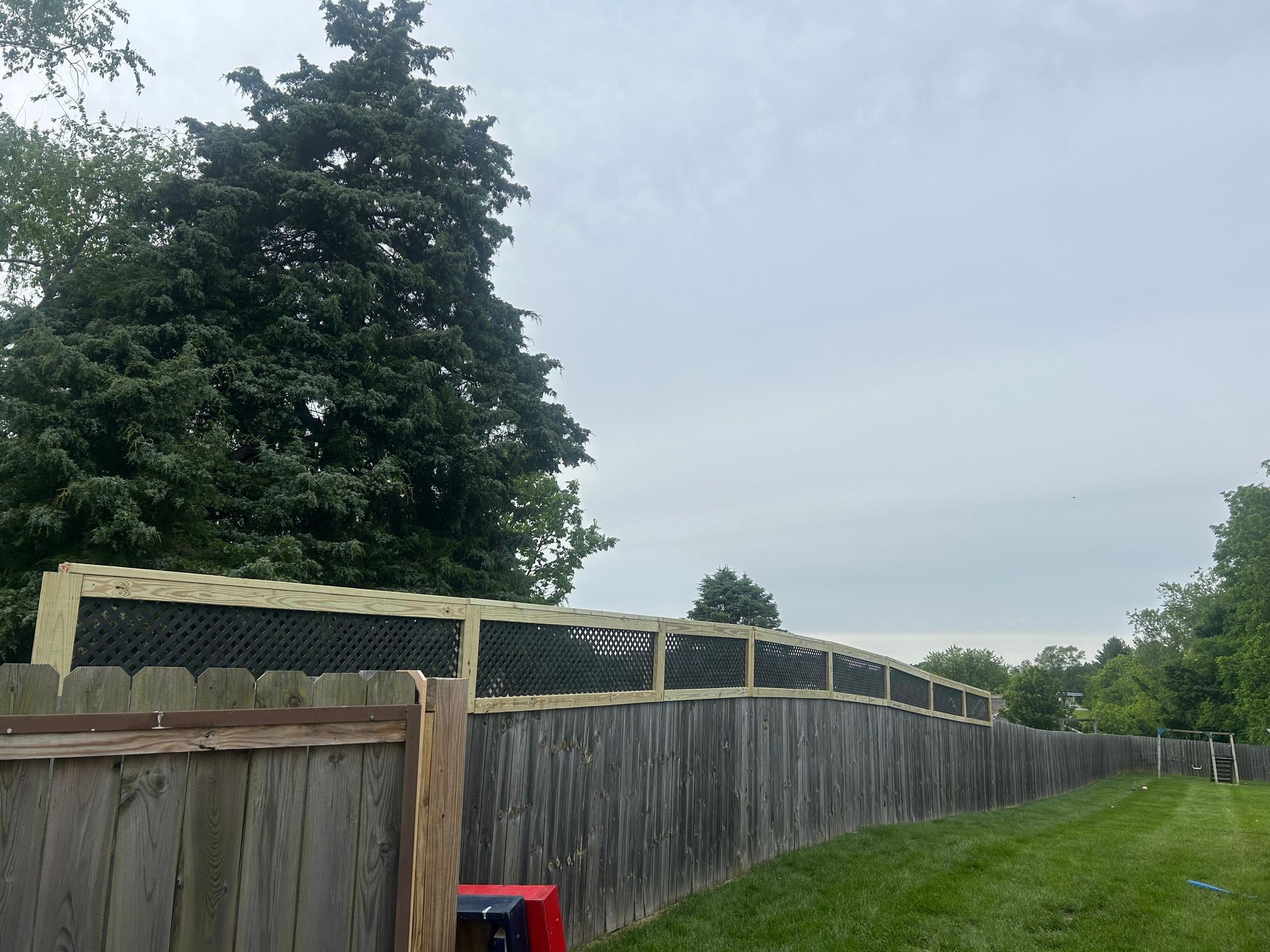 A wooden fence with a new lattice top extension running along a grassy yard under a cloudy sky.
