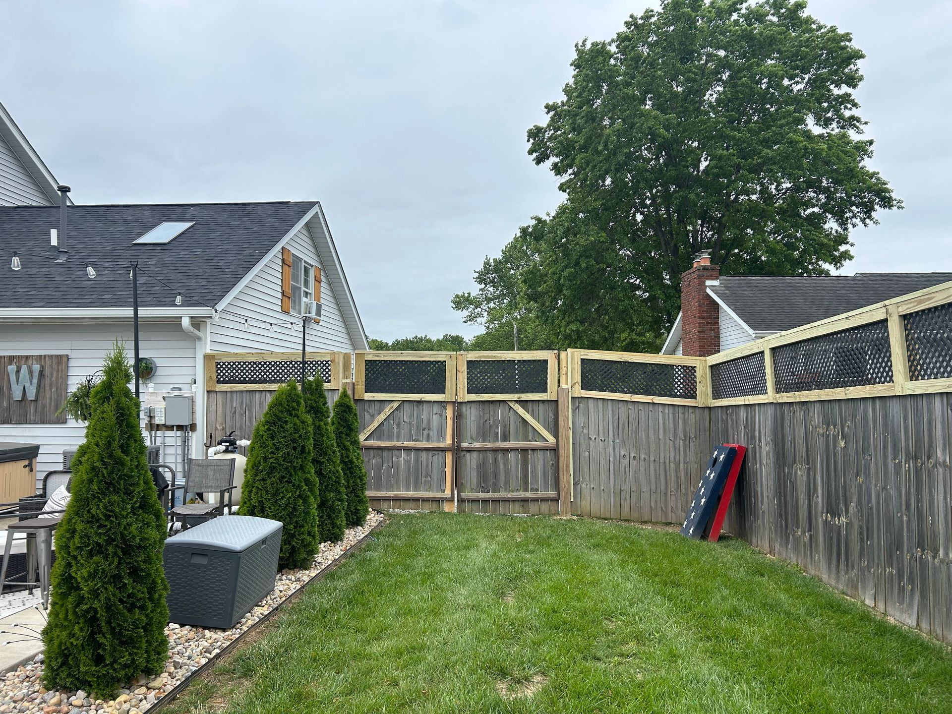 A small grassy backyard with three evergreen shrubs, a gray fence with decorative black panels, and a house exterior.