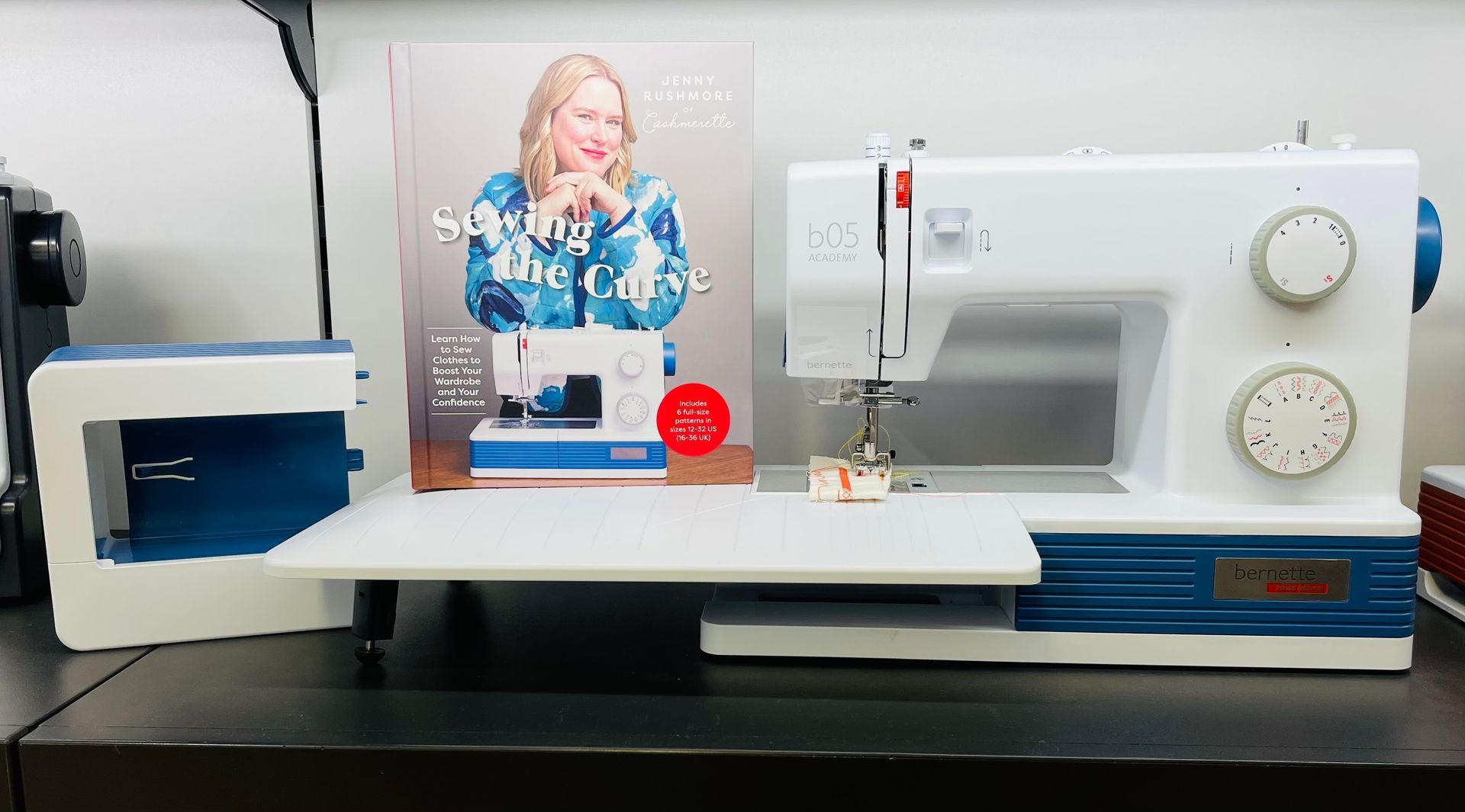 White and blue sewing machine with accessories on a dark table, a book with a woman's image in background.