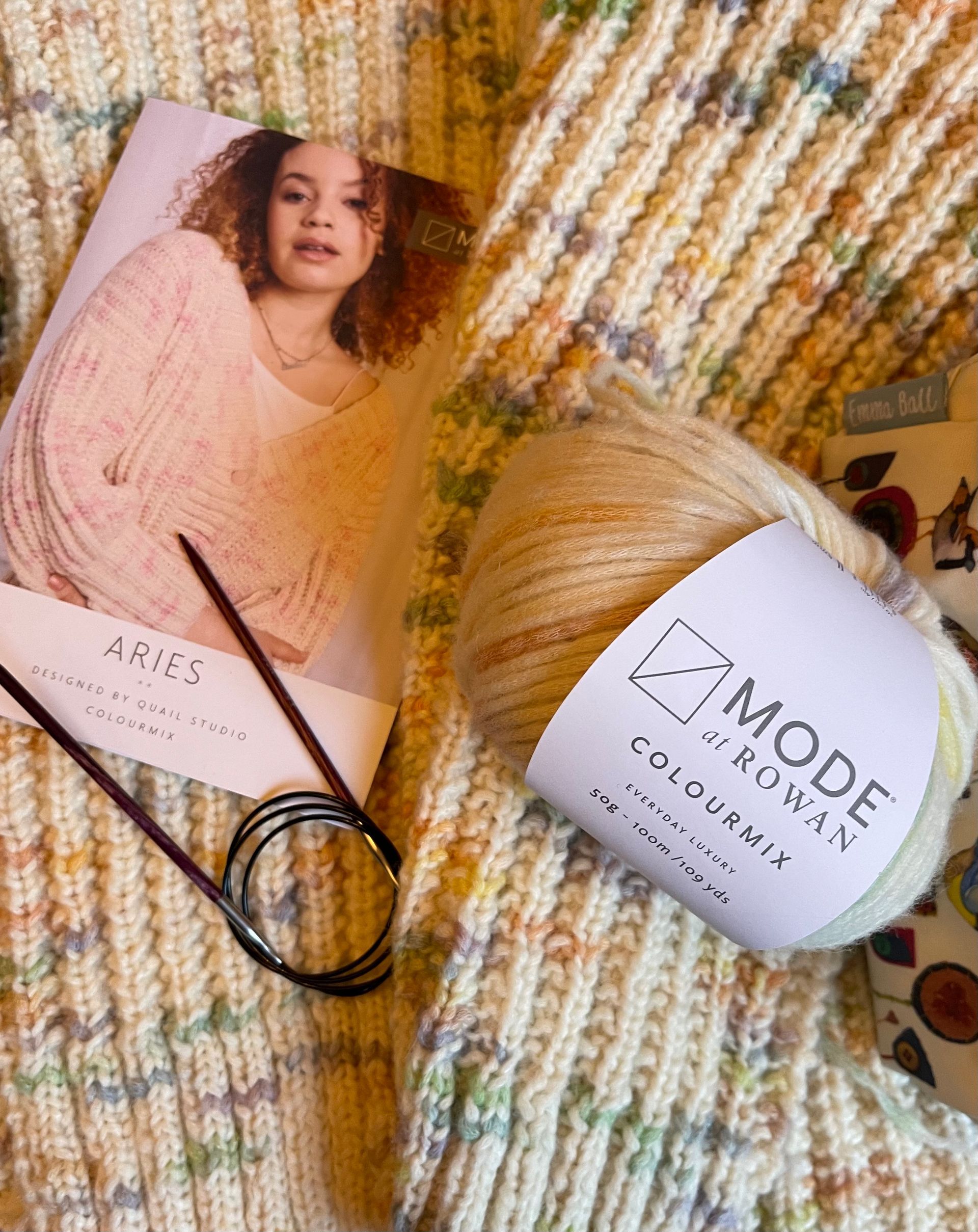 Magazine and yarn skein on a textured woven surface, with a knit project and black crochet hook.