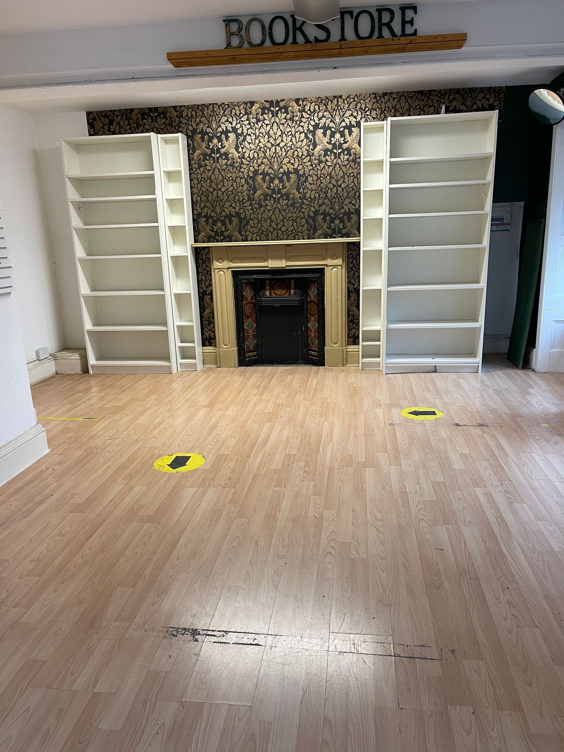 Empty bookstore room with white shelves, a fireplace, and yellow floor markers on a wood floor.