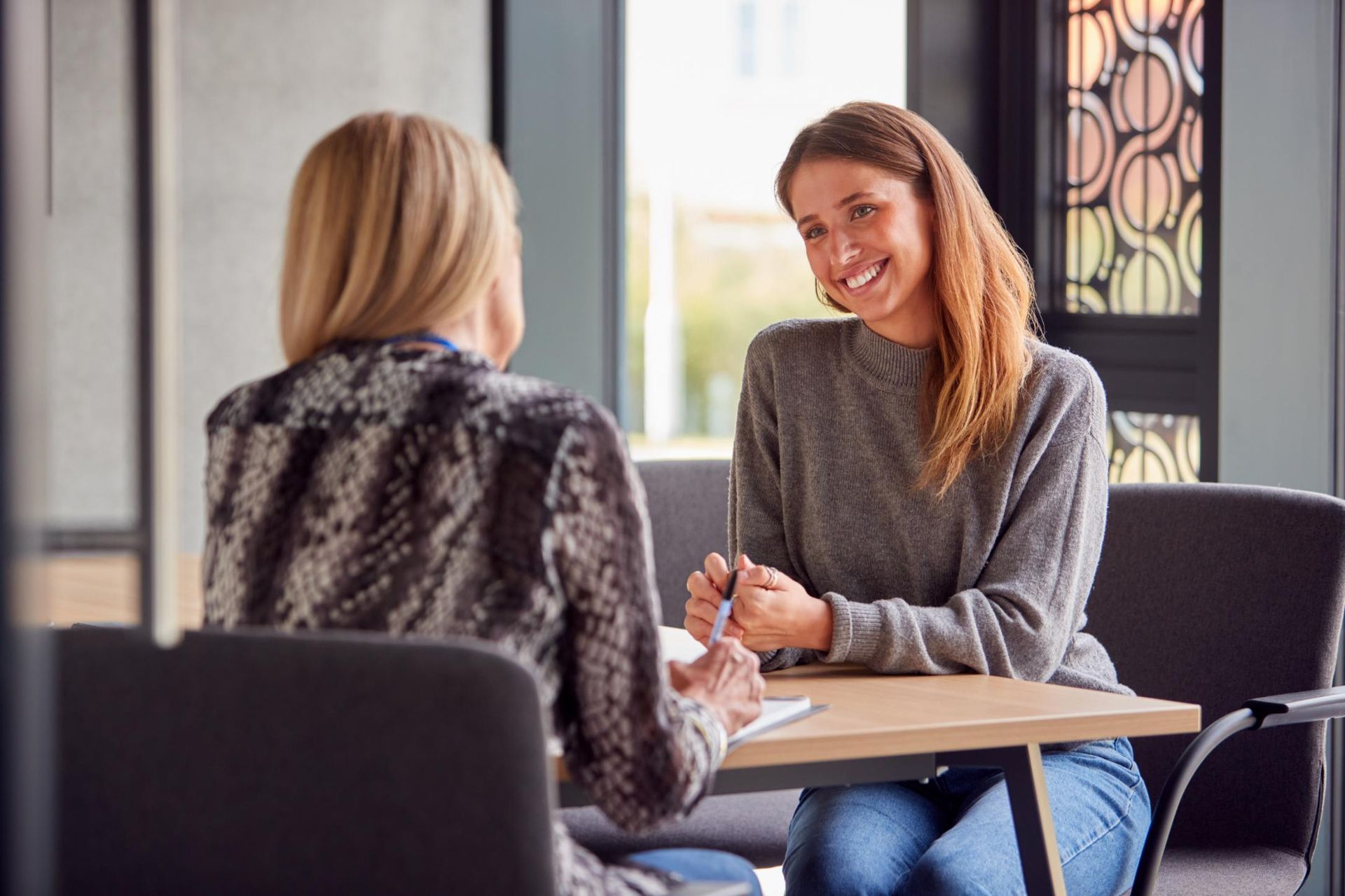 Two women seated at a table, one smiling, other facing away. Light-filled setting.