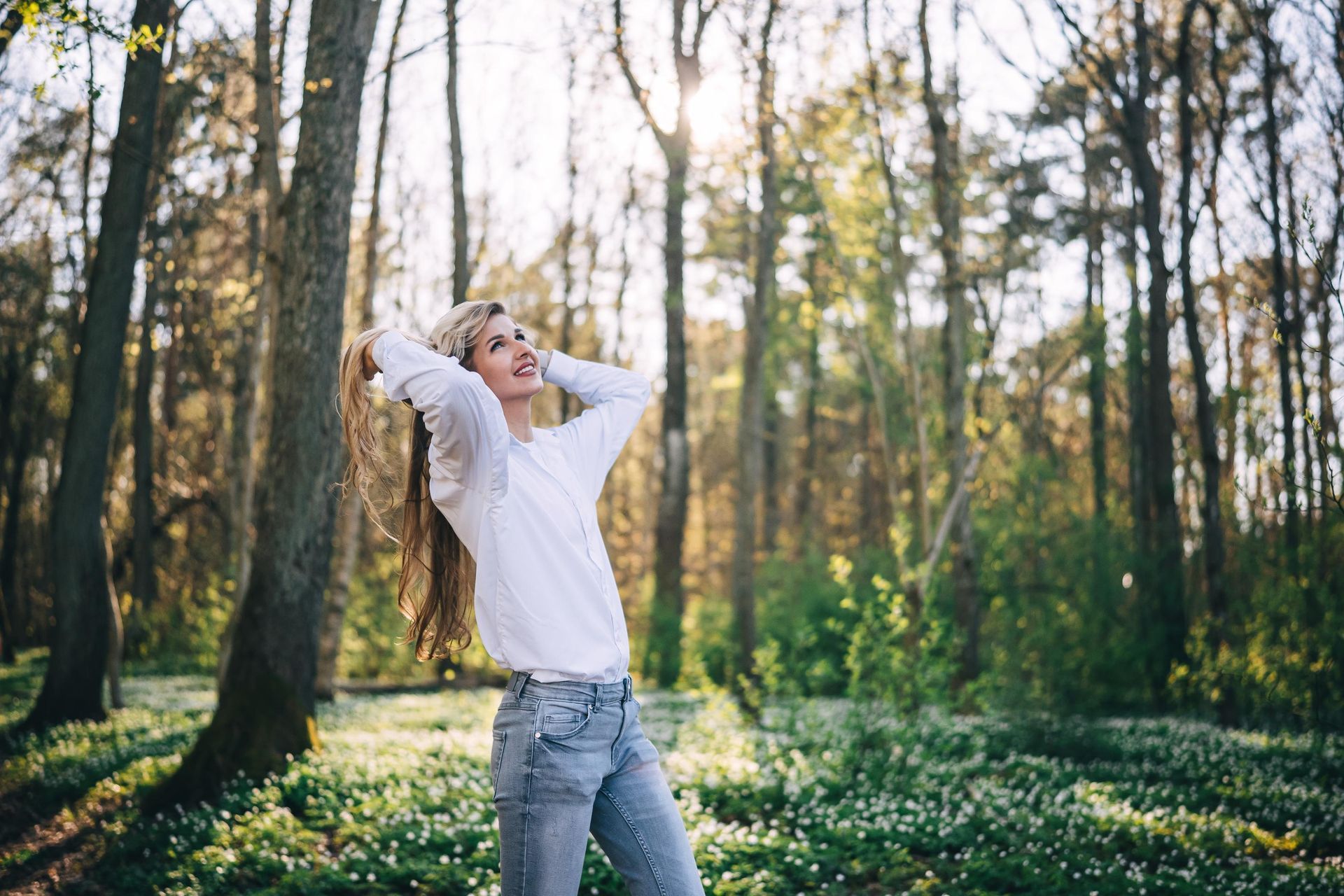 Woman with long blonde hair, smiling, arms raised in a forest. Sunlight streams through trees.