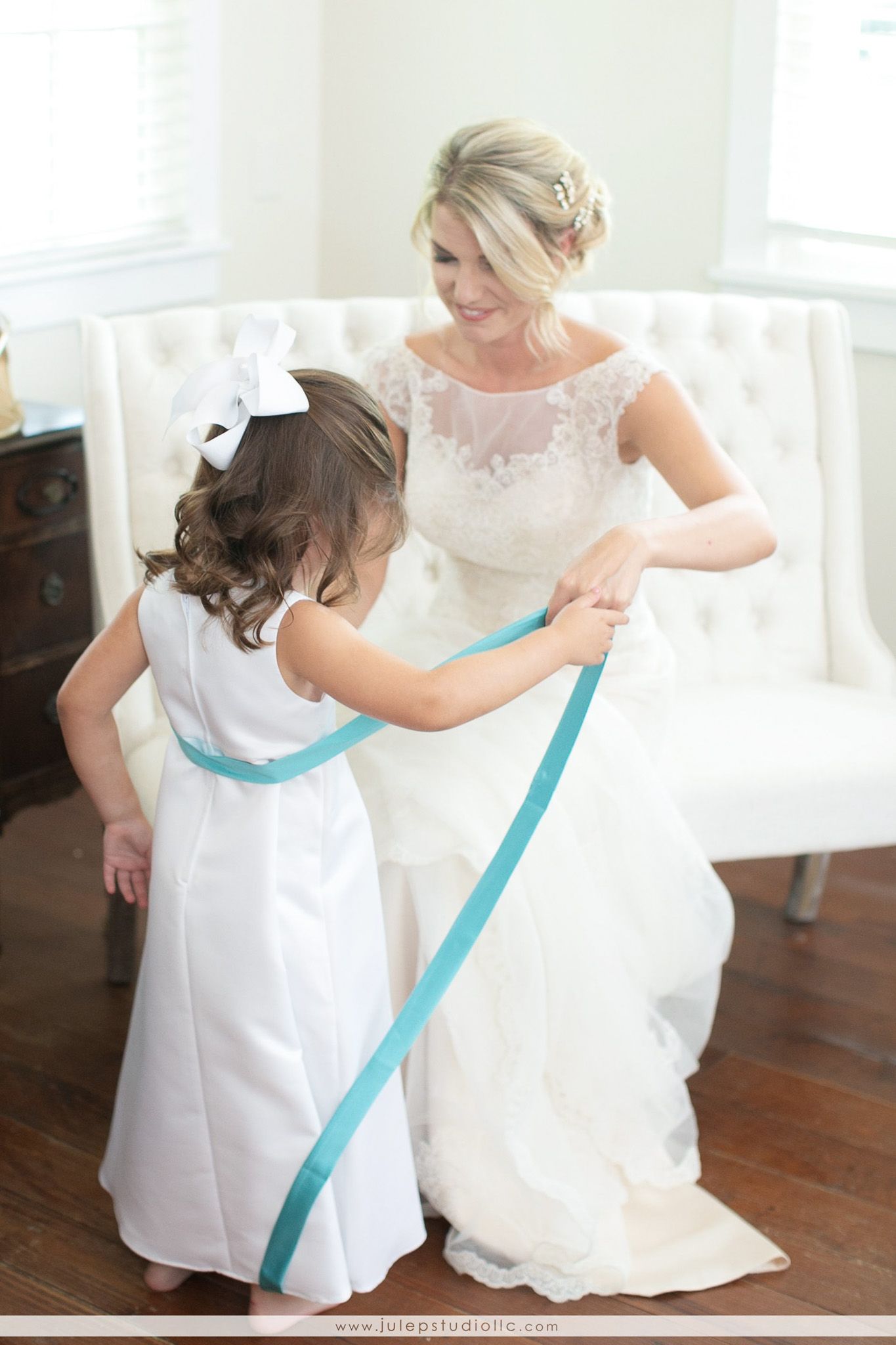 Bride sitting, child with ribbon; indoor setting, light colors.