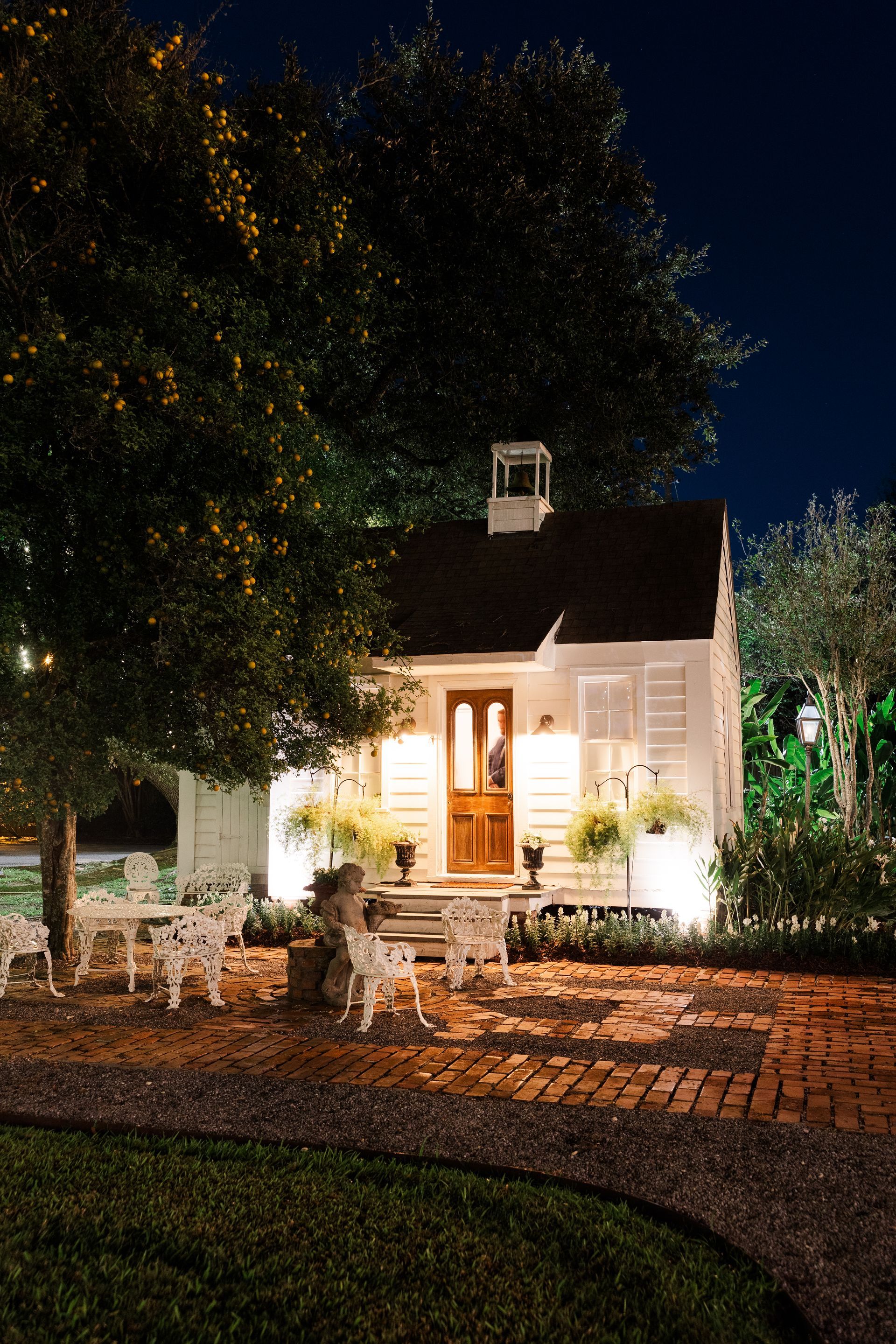 White cottage at night, lit from below; brick pathway, outdoor seating, and lush trees surround.