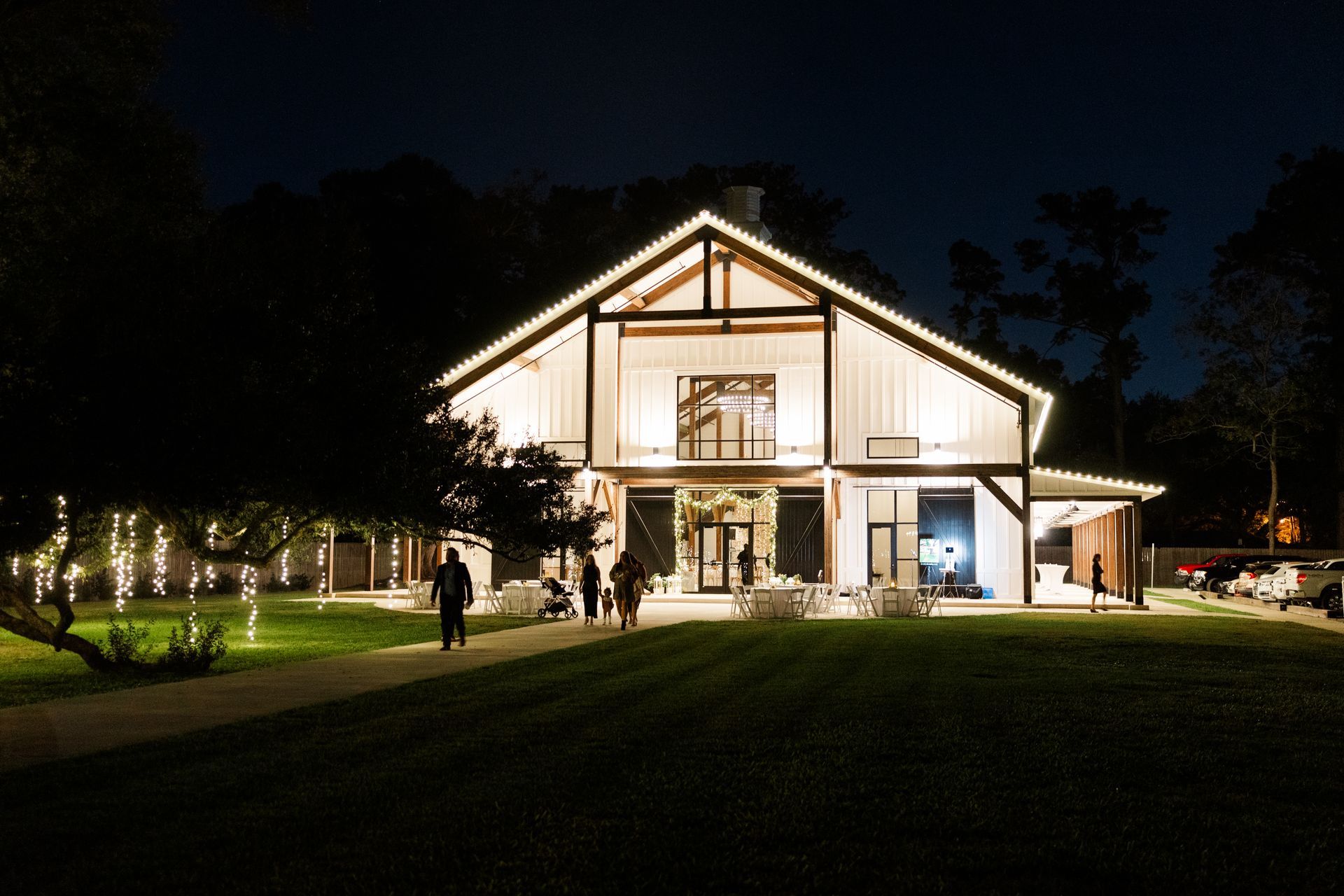 Night view of a white barn lit with string lights. People walk on the lawn towards the entrance.