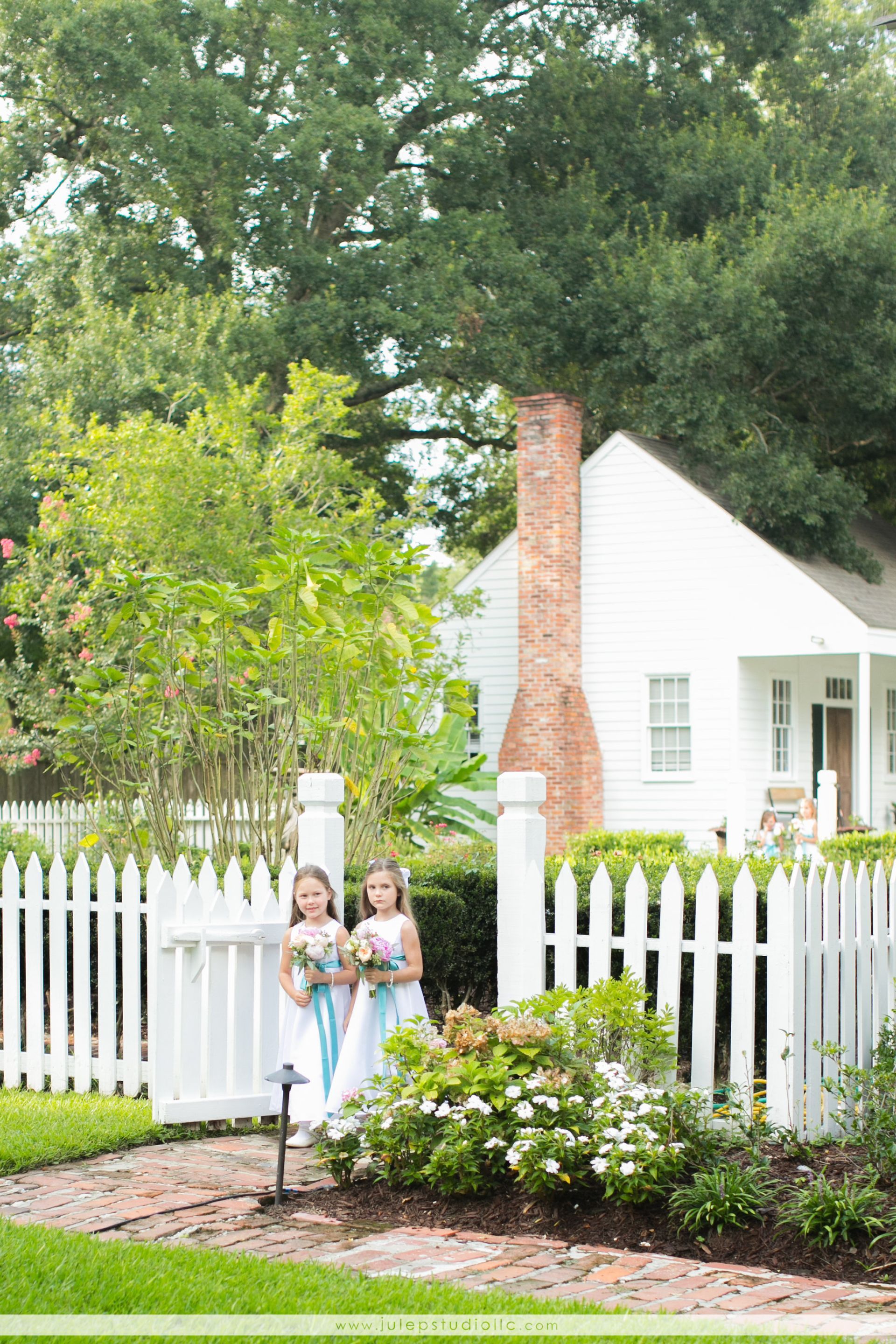 Two girls in white dresses holding bouquets stand by a white picket fence in front of a white house.