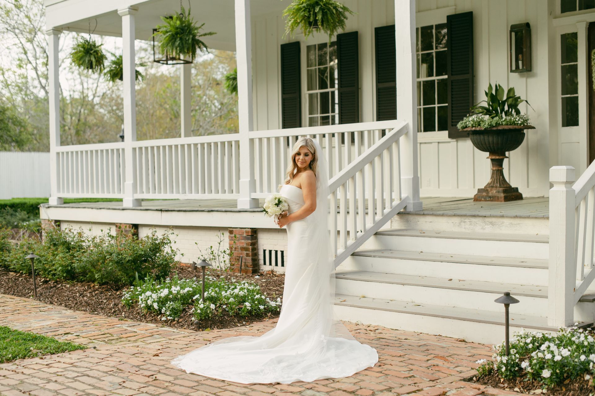 Bride in a white gown stands on porch steps, holding flowers. White house with shutters, greenery.