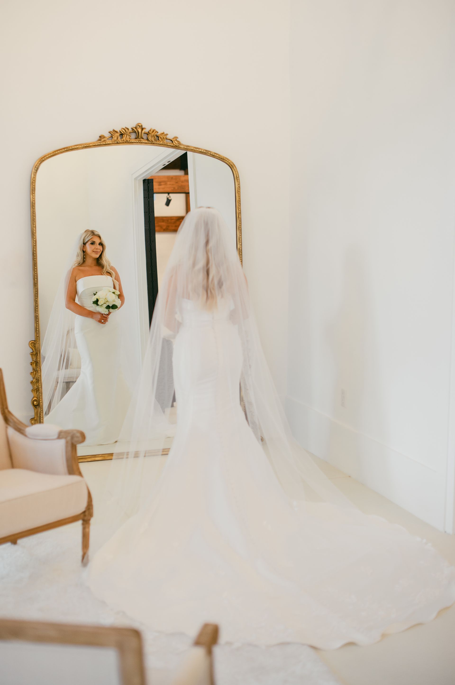 Bride in a white gown and veil looks at herself in a gold-framed mirror.