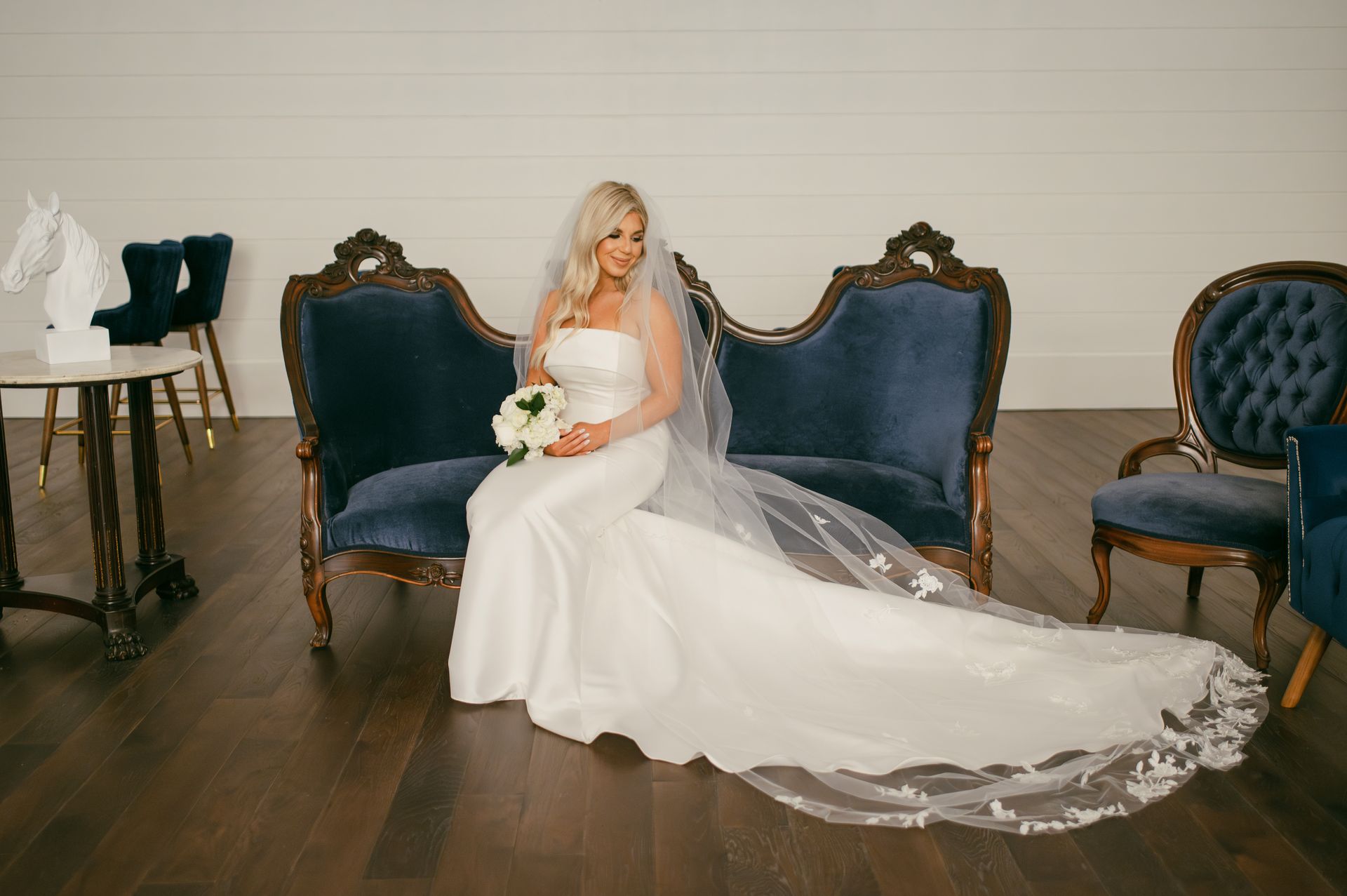 Bride in white gown with long veil sits on blue velvet antique sofa, holding bouquet.