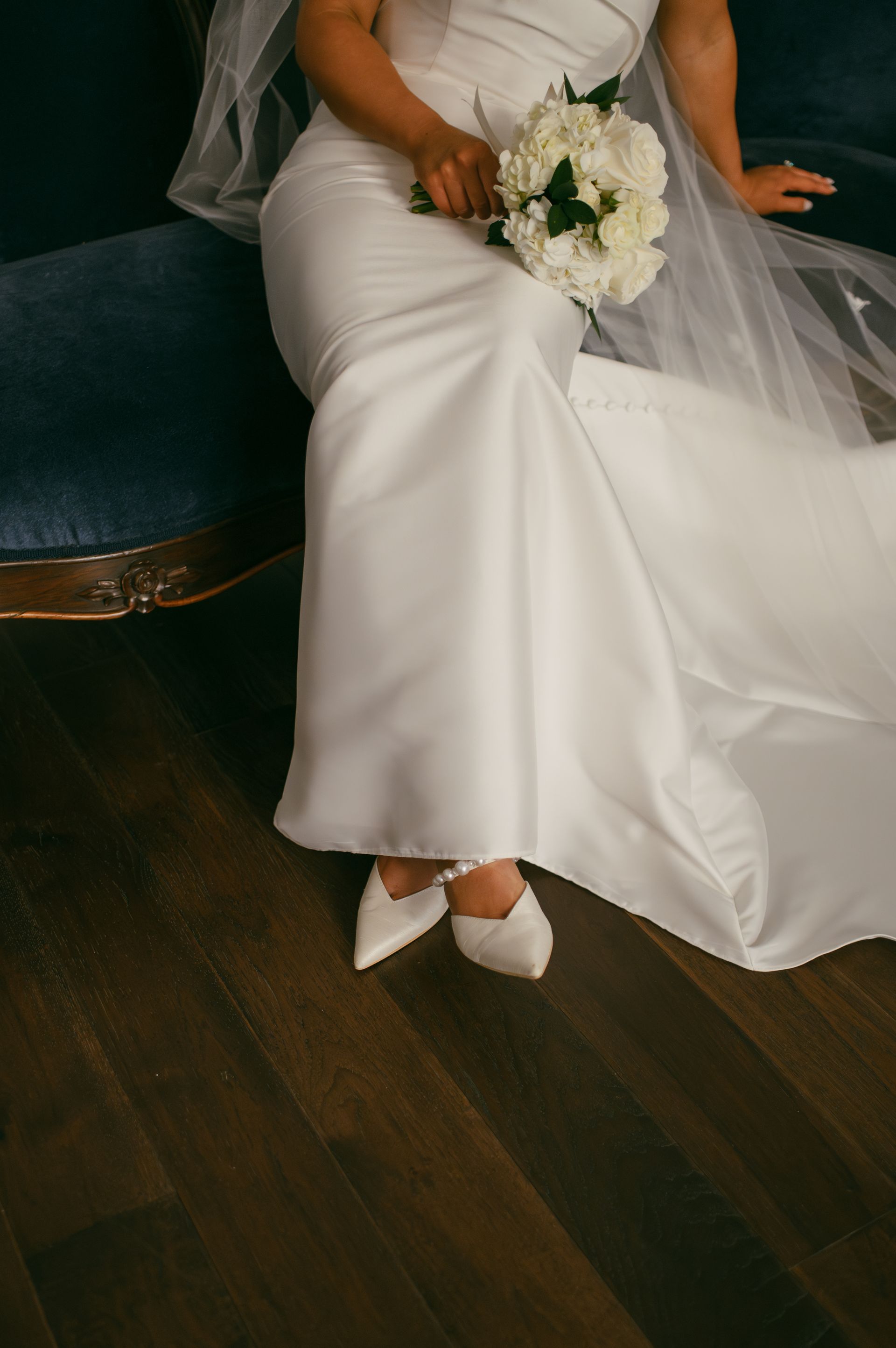 Bride in white gown holding bouquet, sitting on blue velvet chair, white shoes, dark wood floor.