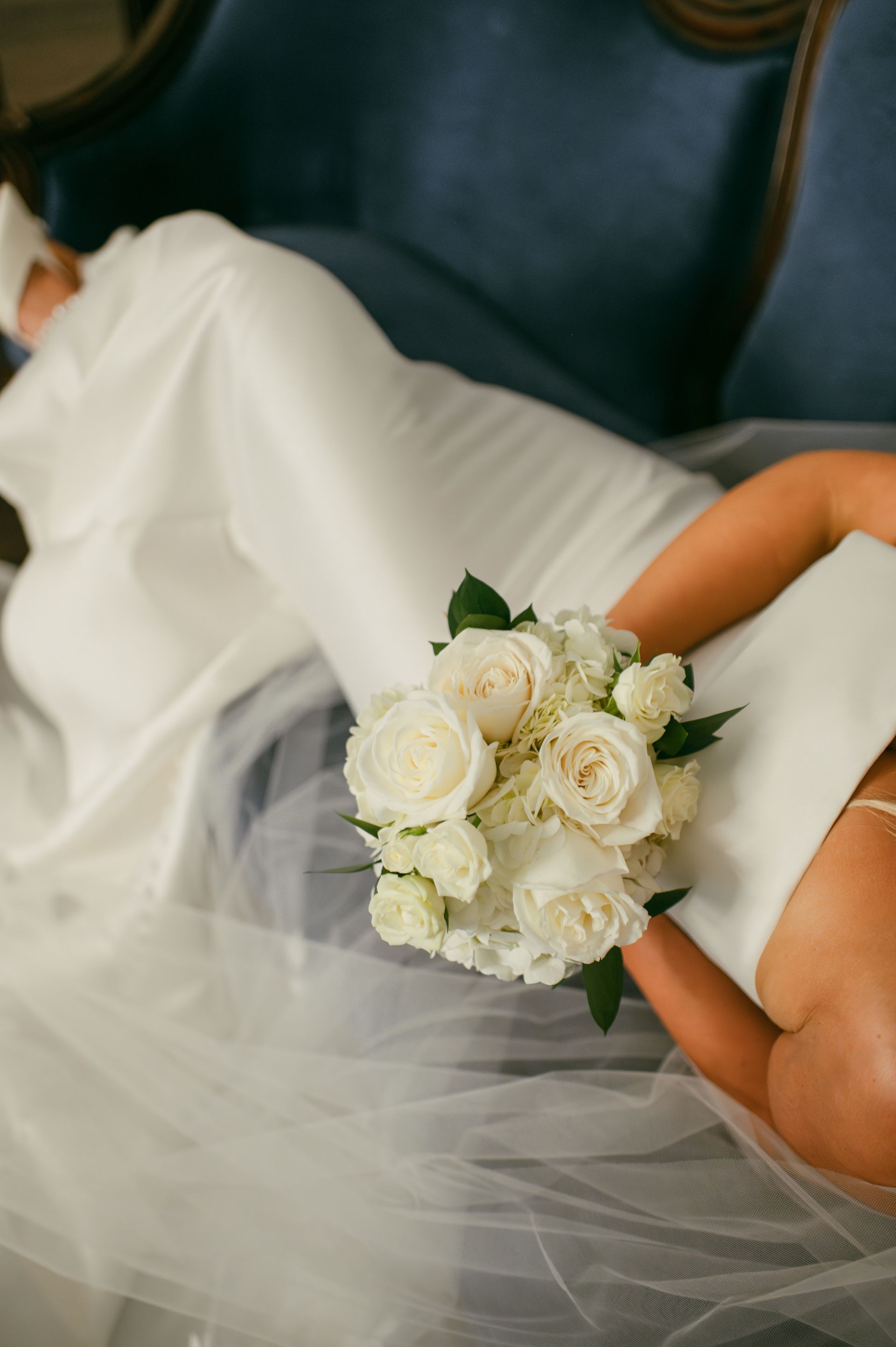 Bride in white dress holding bouquet of white flowers, reclining on a blue velvet sofa.