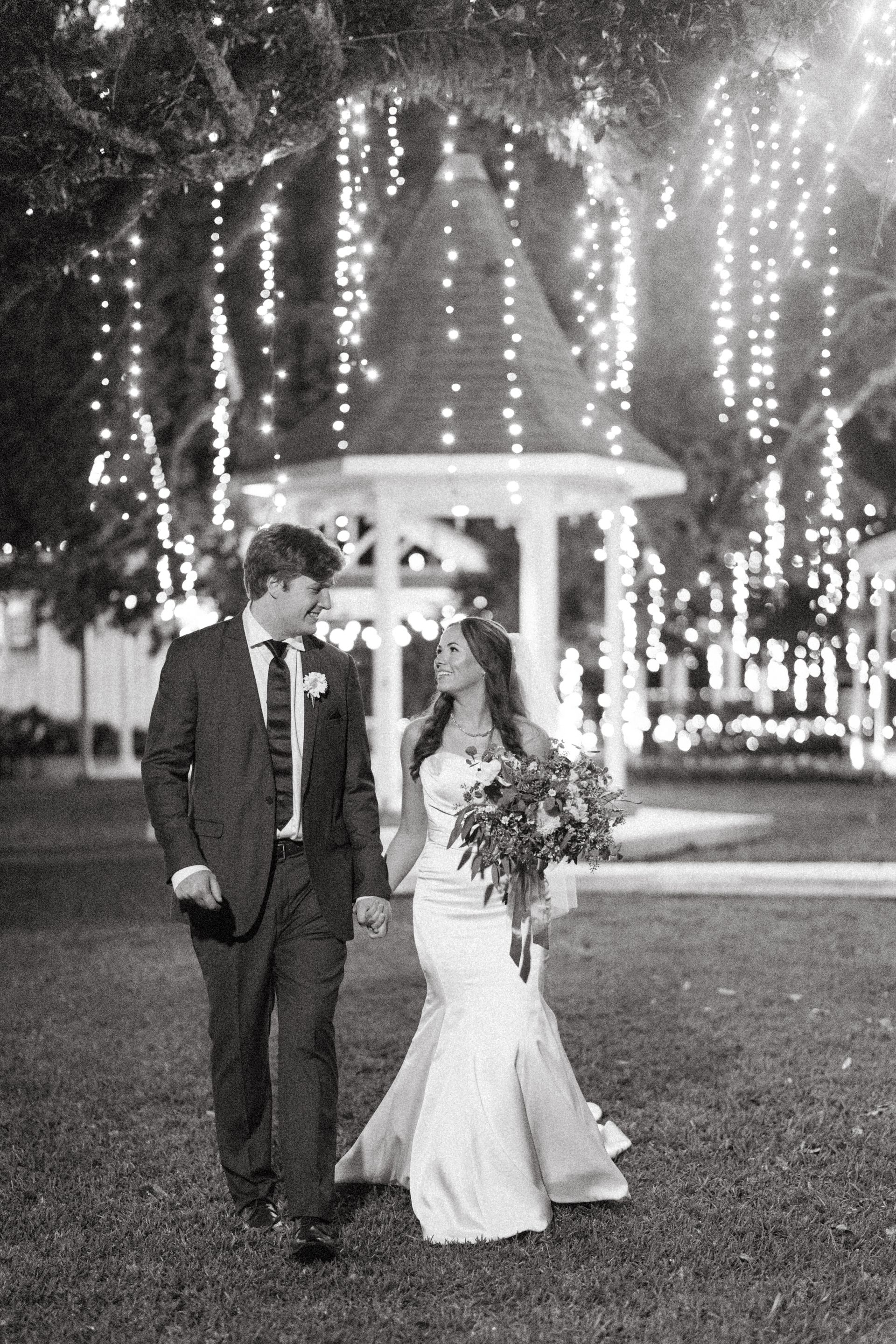 Newlyweds hold hands and walk at night, under strings of lights and near a gazebo.