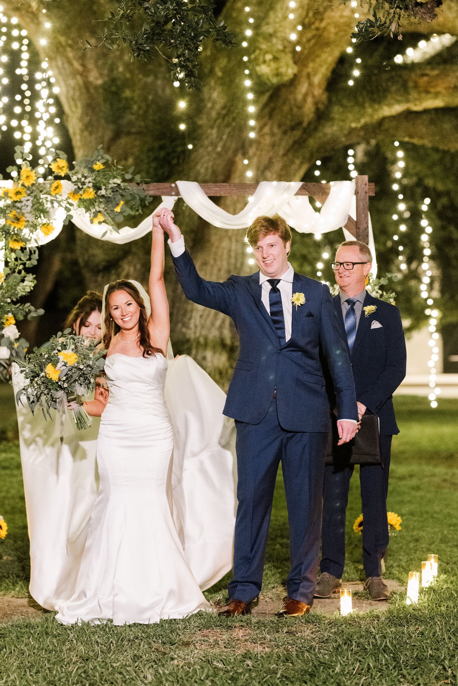 Newlyweds raise arms under a decorated arch; outdoor wedding.