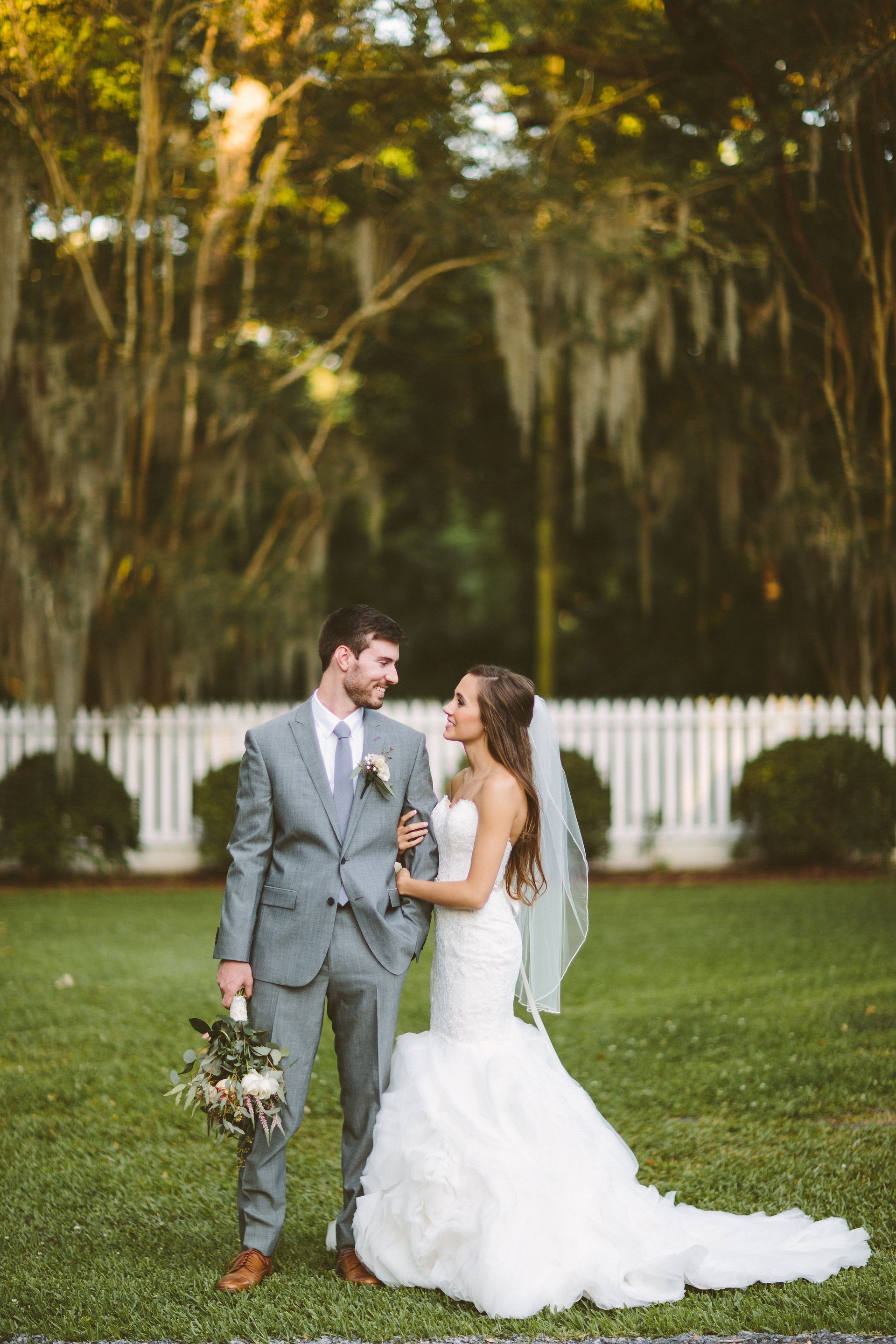 Bride and groom pose on grass in front of a white picket fence, looking at each other, holding hands.