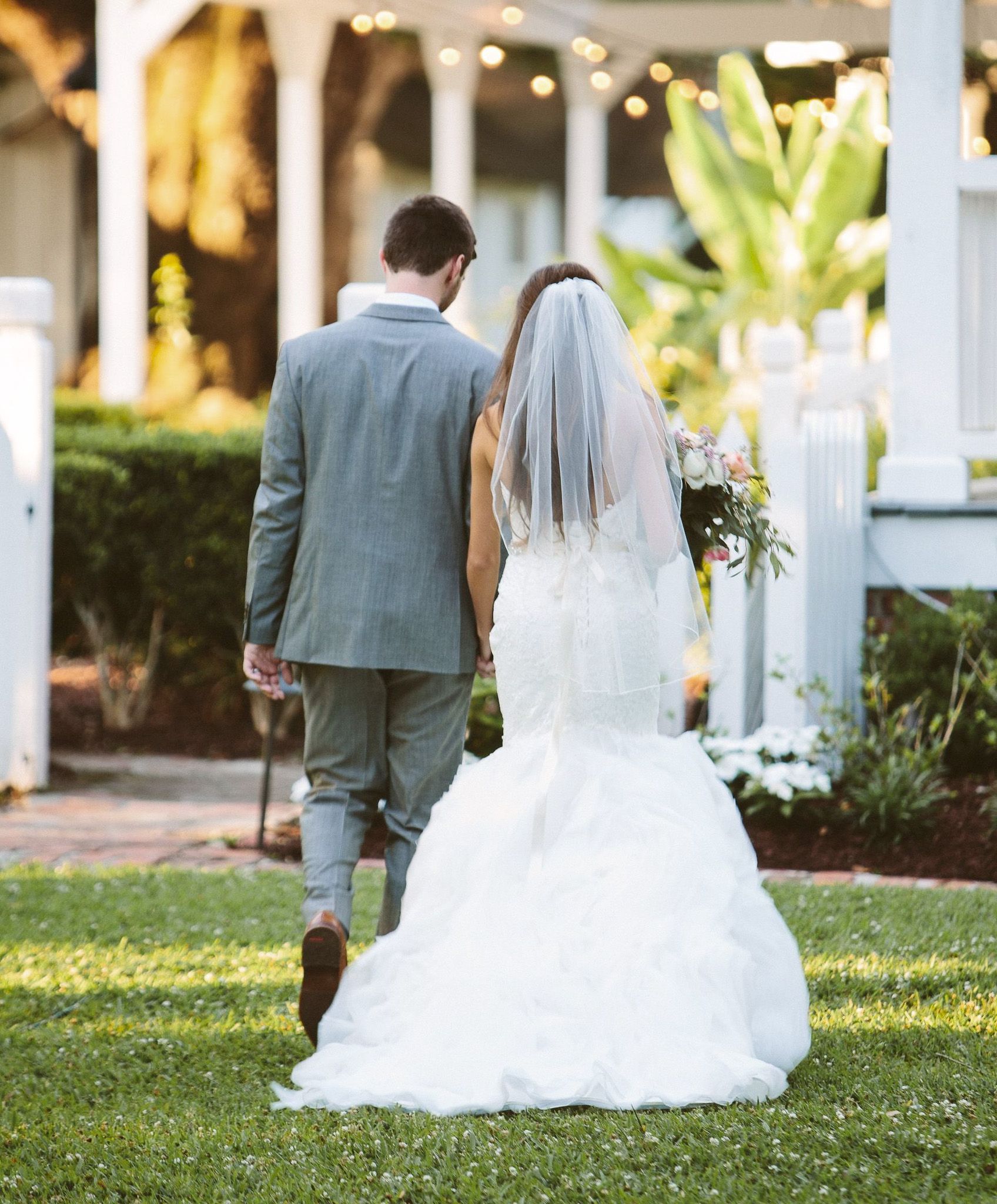 Bride and groom walking on grass, holding hands. Bride wears white lace dress, veil, and carries flowers. Groom in gray suit.