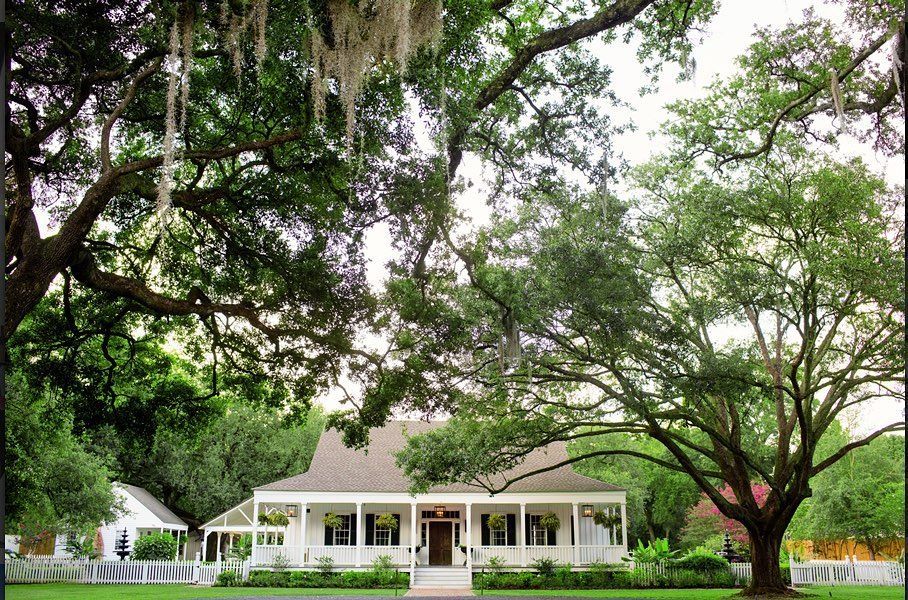 White house with porch, surrounded by large, leafy trees and a white picket fence.