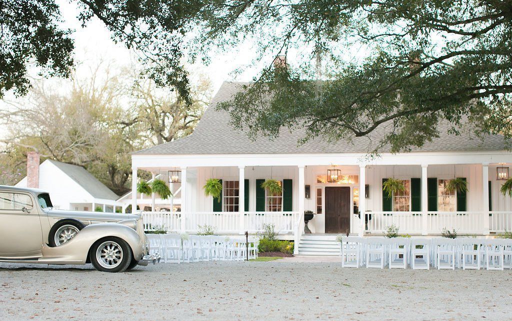 White house with porch, wedding ceremony setup. Classic car parked in front.