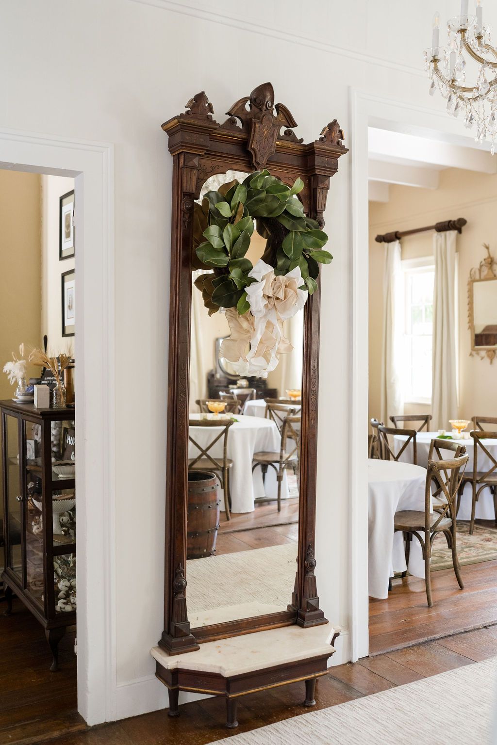 Tall antique mirror with carved frame and wreath, reflecting a dining room.