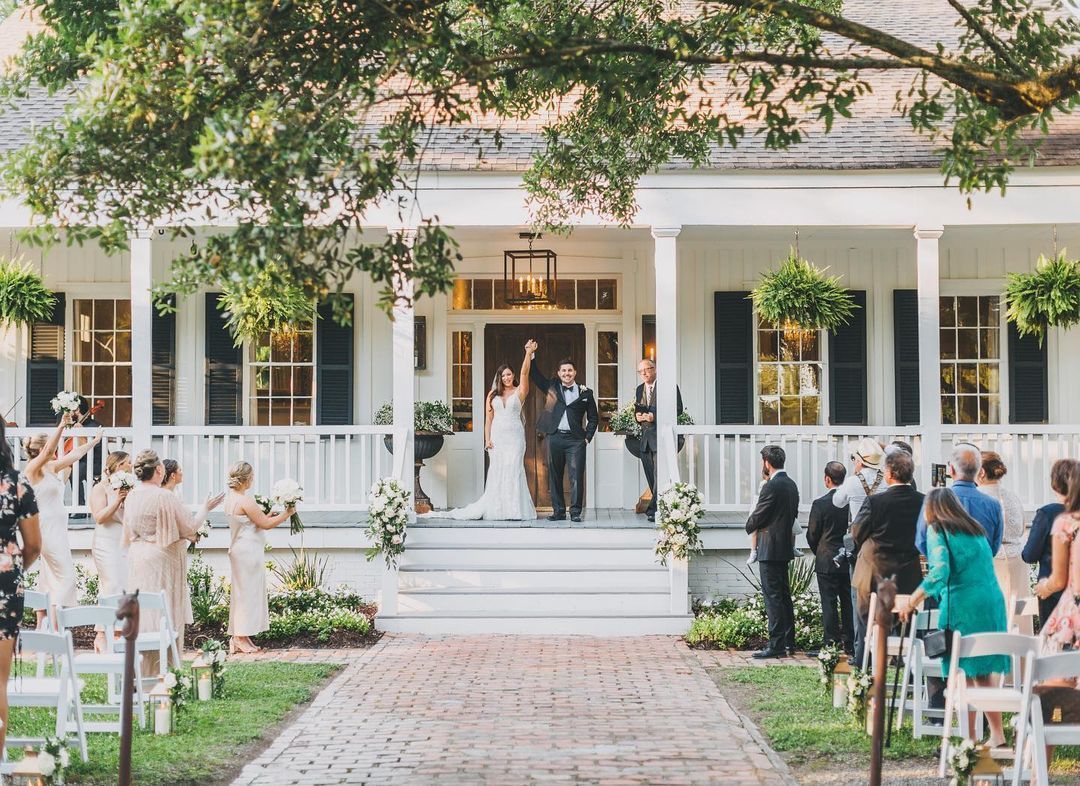 Newlyweds celebrate on a porch, surrounded by wedding guests. Ceremony outside a white house.