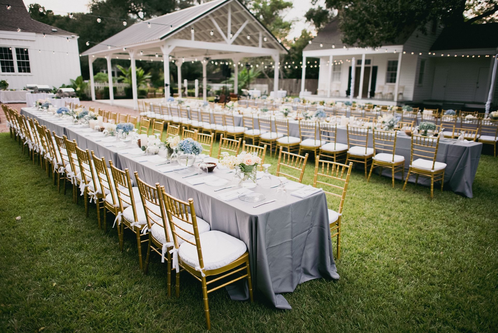 Long tables set for a formal outdoor event, with gold chairs, gray tablecloths, and floral centerpieces under a white gazebo.