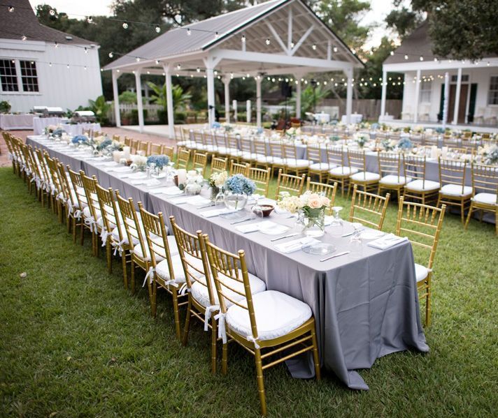 Long outdoor dining tables set for a celebration, with gold chairs, gray tablecloths, and floral centerpieces.