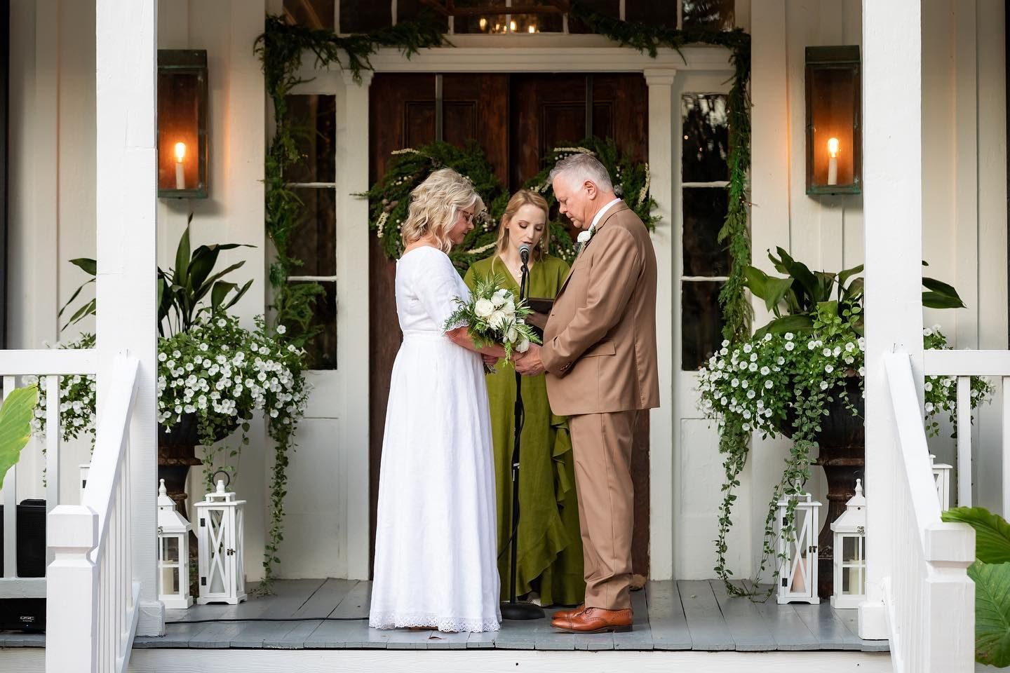 A couple exchanging vows on a porch, officiated by a woman. Greenery and lanterns decorate the space.