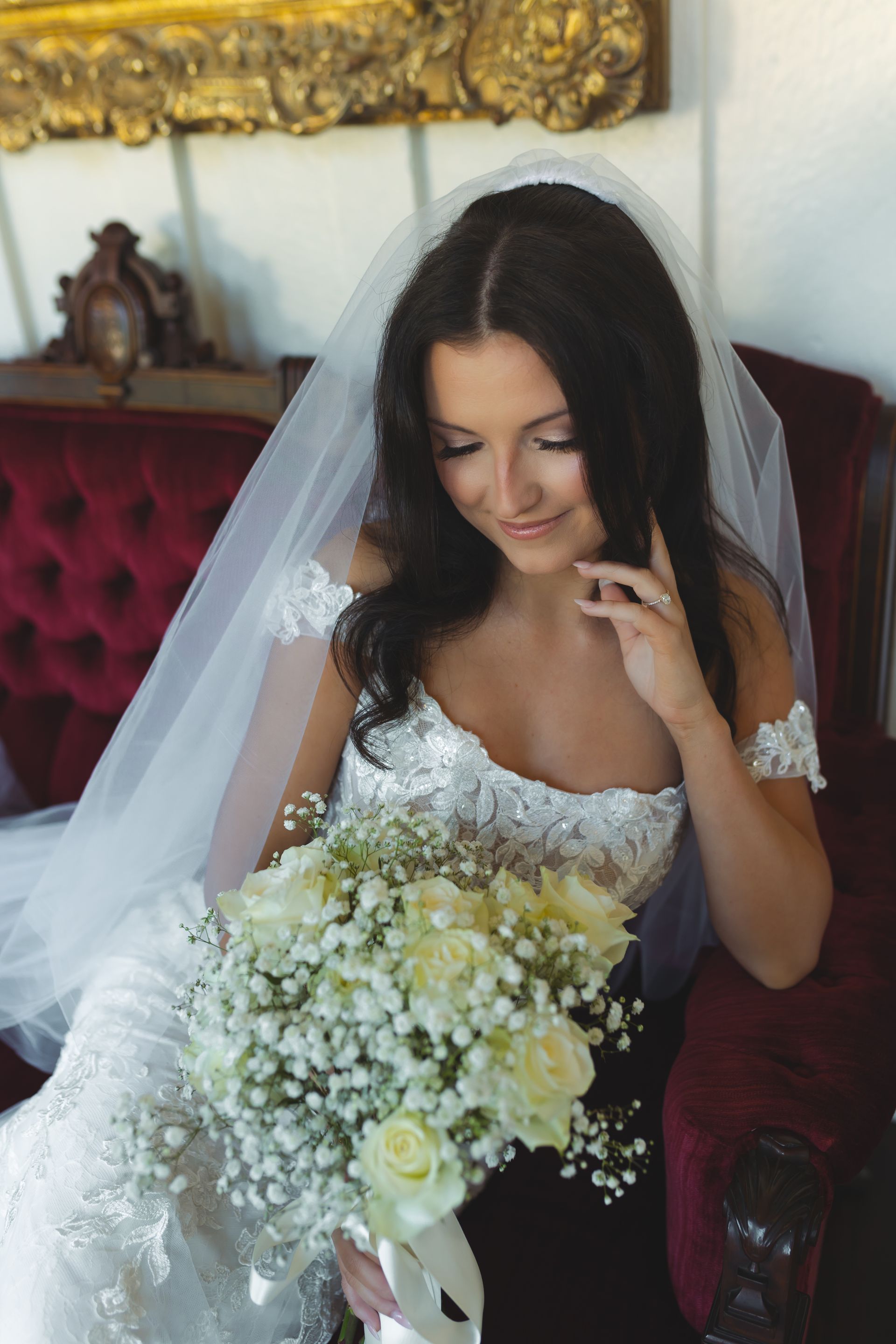 Bride in white wedding dress, holding bouquet, looking down, sitting on red velvet chair.
