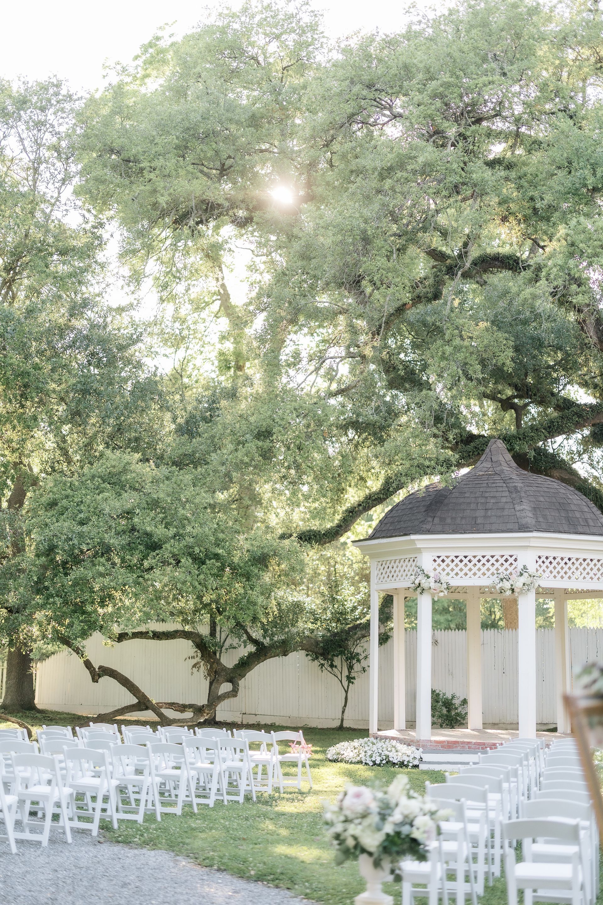 Wedding ceremony setup with white chairs, gazebo, and large trees.
