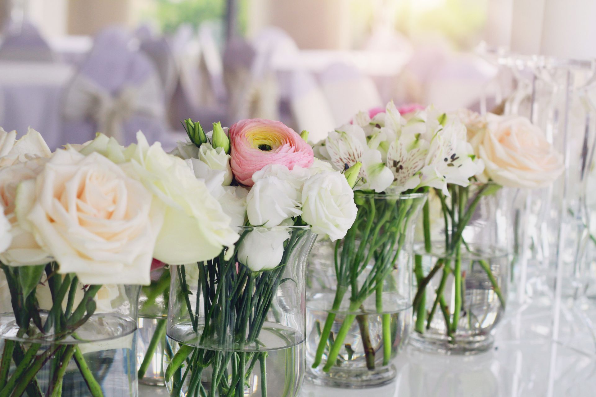 Vases of pastel flowers, including roses, on a white table. Blurred background with more tables.