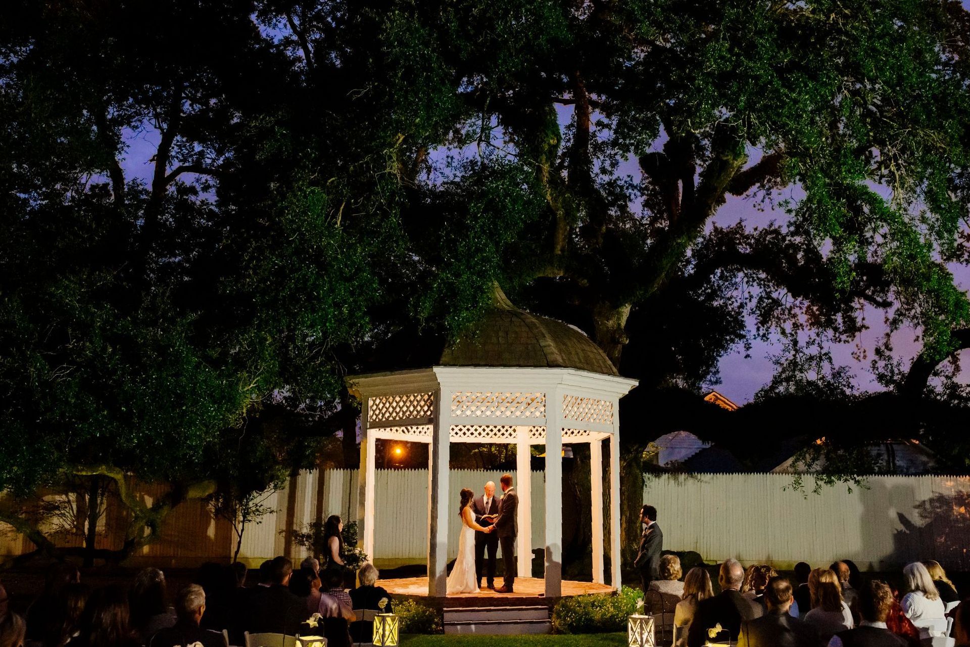 Wedding ceremony under a large tree; couple in a gazebo, guests seated in front.
