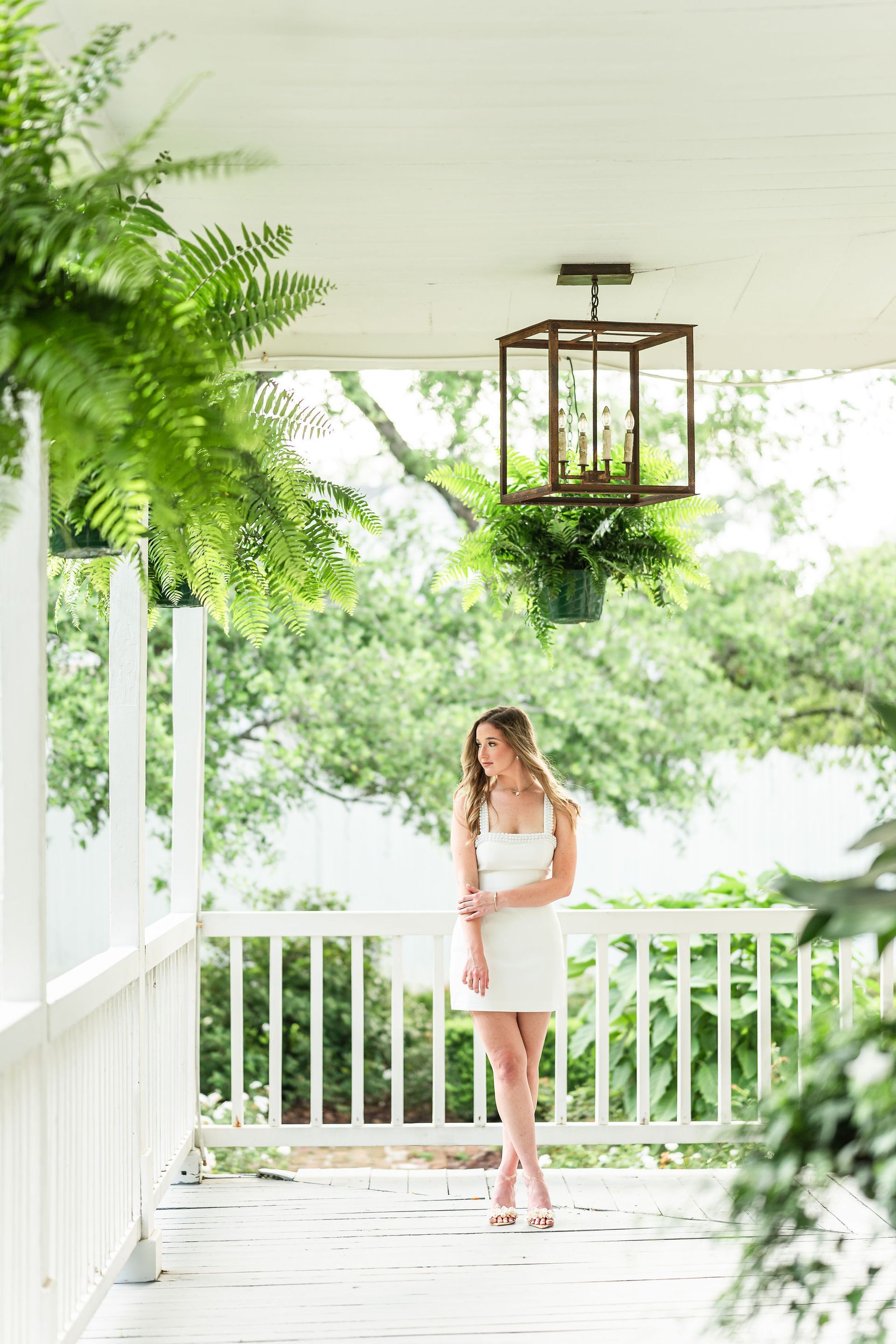 Woman in a white dress stands on a porch with ferns, looking to the side. Wooden light fixture hangs overhead.