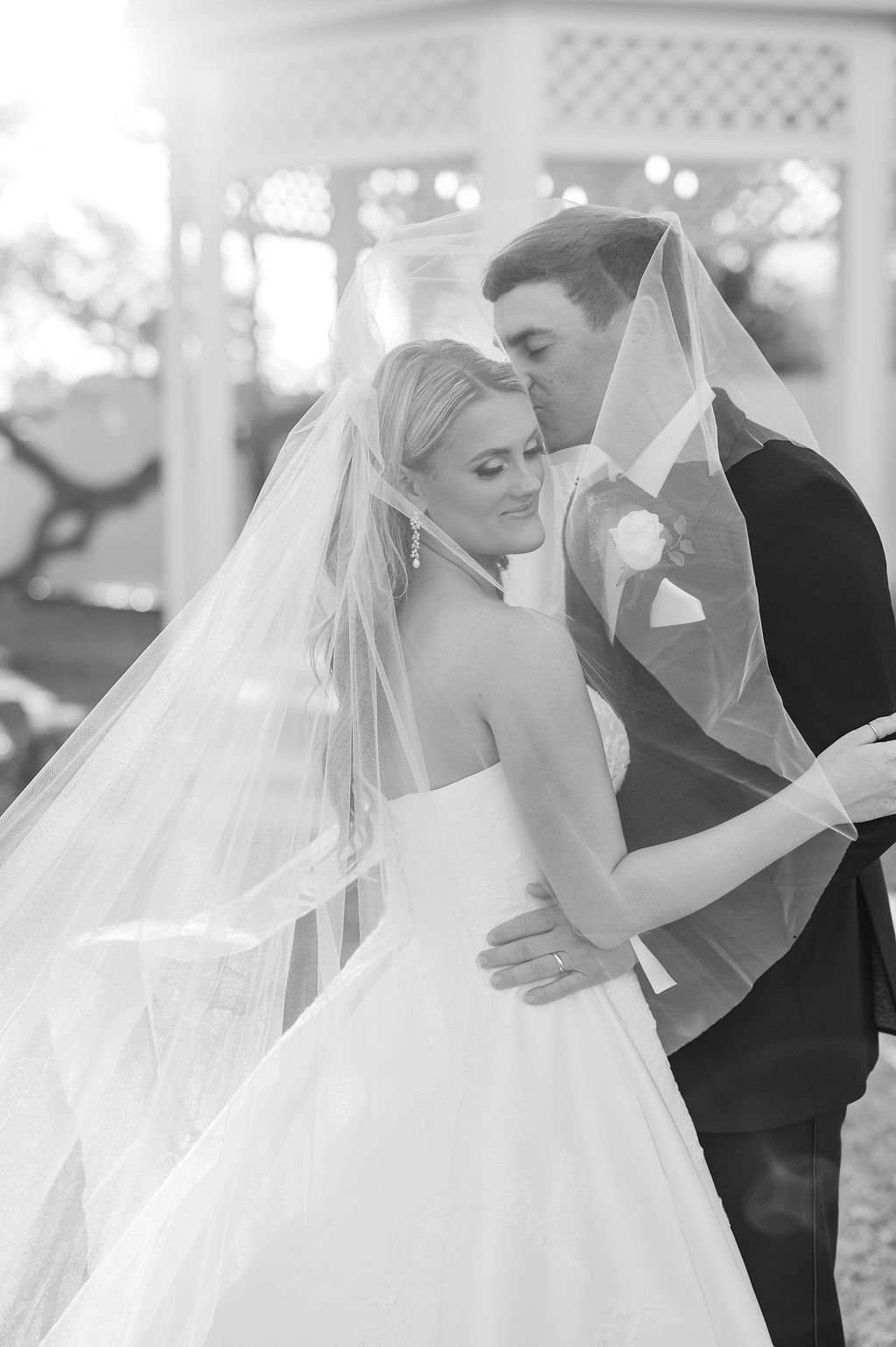 Bride and groom share a tender moment. The groom kisses the bride's forehead, under her veil, near a gazebo.