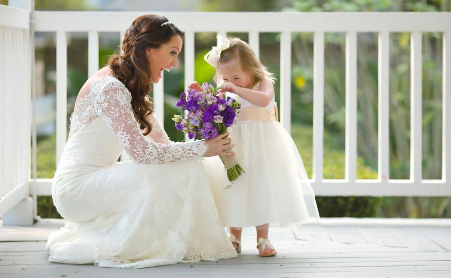Bride in a white gown and flower girl in a white dress with purple flowers on a porch.